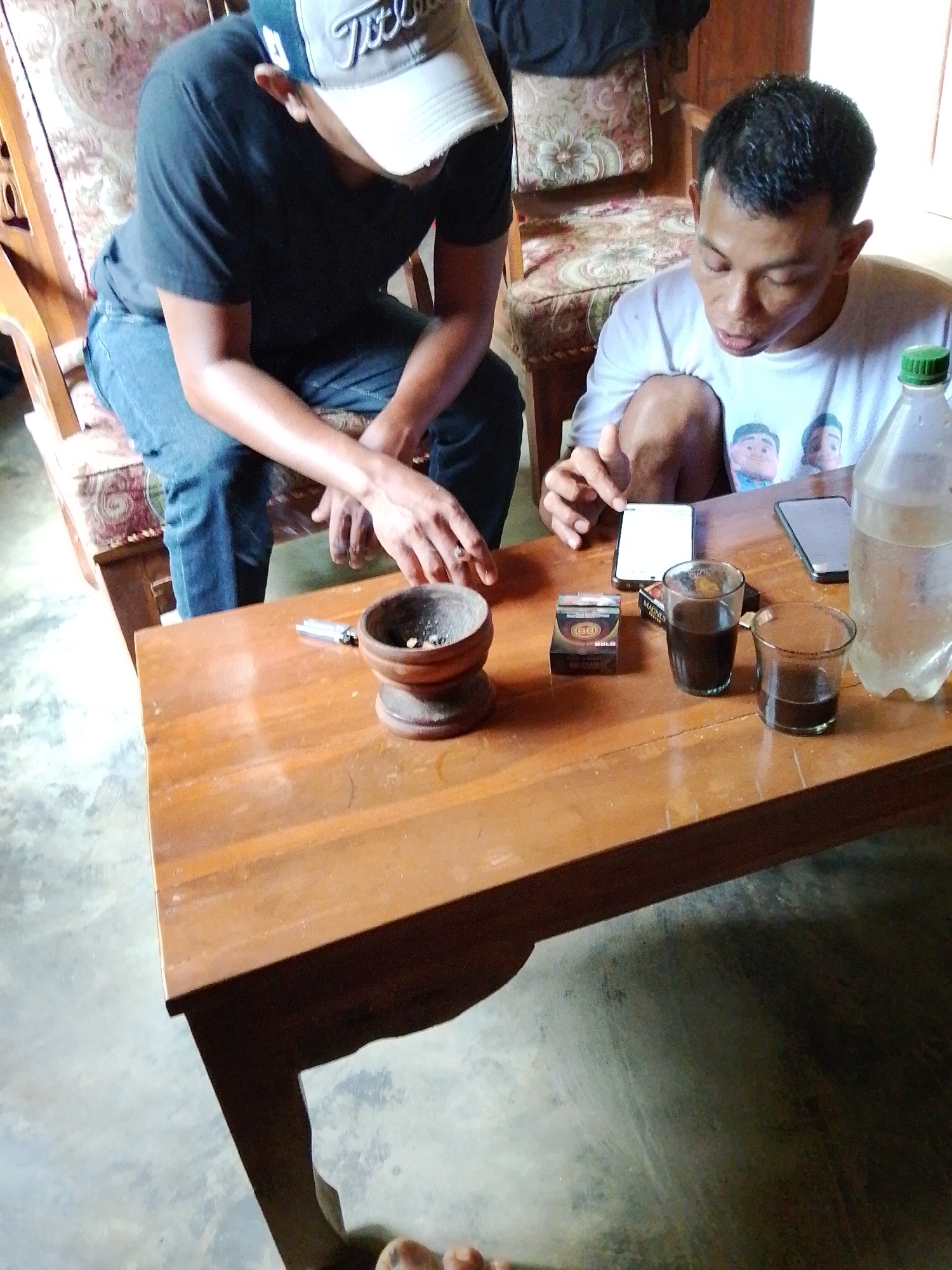 The image depicts two individuals seated at a wooden table in what appears to be a residential setting in Madukoro, Indonesia. On the table, a pack of cigarettes, two glasses of dark liquid, a lighter, and a small wooden bowl are visible. A plastic bottle of water stands to the right. One person, wearing a black t-shirt and a baseball cap, is on the left, and the other, in a white t-shirt, is on the right, looking down at the table. The environment is casual, with upholstered chairs in the background.