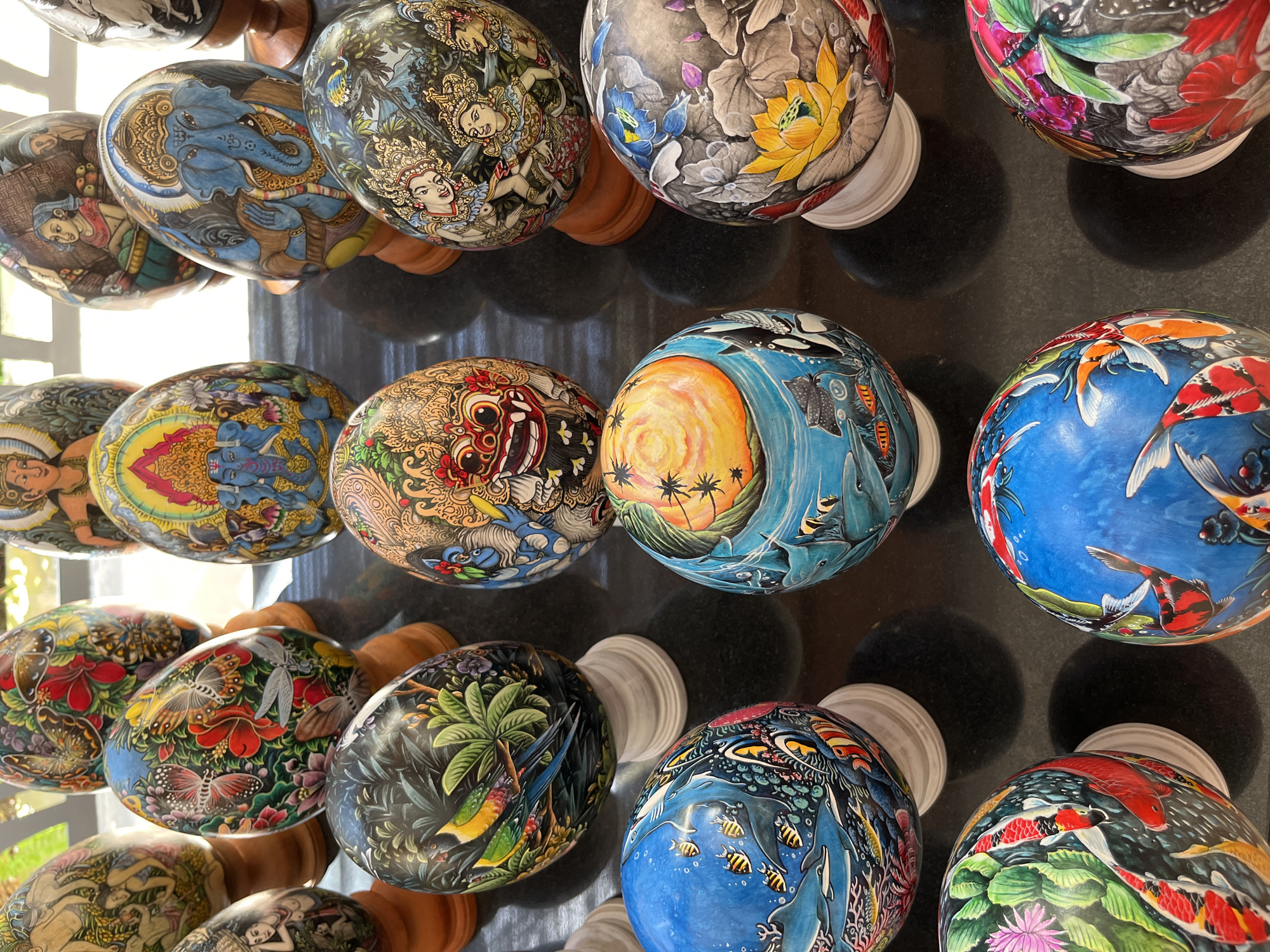 In Kuta Selatan, Indonesia, a collection of intricately painted decorative eggs is displayed on a dark, reflective surface. The eggs, each unique, are presented on small wooden or white stands, suggesting they are for sale or exhibition.
The eggs are adorned with a variety of vibrant and detailed artwork, predominantly featuring mythological figures, animals, and floral motifs in a style reminiscent of Balinese art. Some eggs depict Hindu deities like Ganesha, with his distinctive elephant head, and other figures in elaborate traditional clothing. There are also eggs painted with colorful birds, tropical foliage, aquatic scenes featuring fish, dolphins, and stingrays, and intricate patterns of flowers and insects. The artwork is characterized by its rich colors, fine lines, and sense of depth.
The lighting appears to be natural, possibly from a window or open area, casting soft shadows on the surface and highlighting the textures and colors of the eggs. The arrangement suggests a marketplace, a souvenir shop, or a handicraft display. No people are visible in the immediate foreground or interacting with the eggs. The background is slightly blurred, but hints of diffused light and possibly architectural elements are present. The overall impression is one of artisanal craftsmanship and cultural artistry.