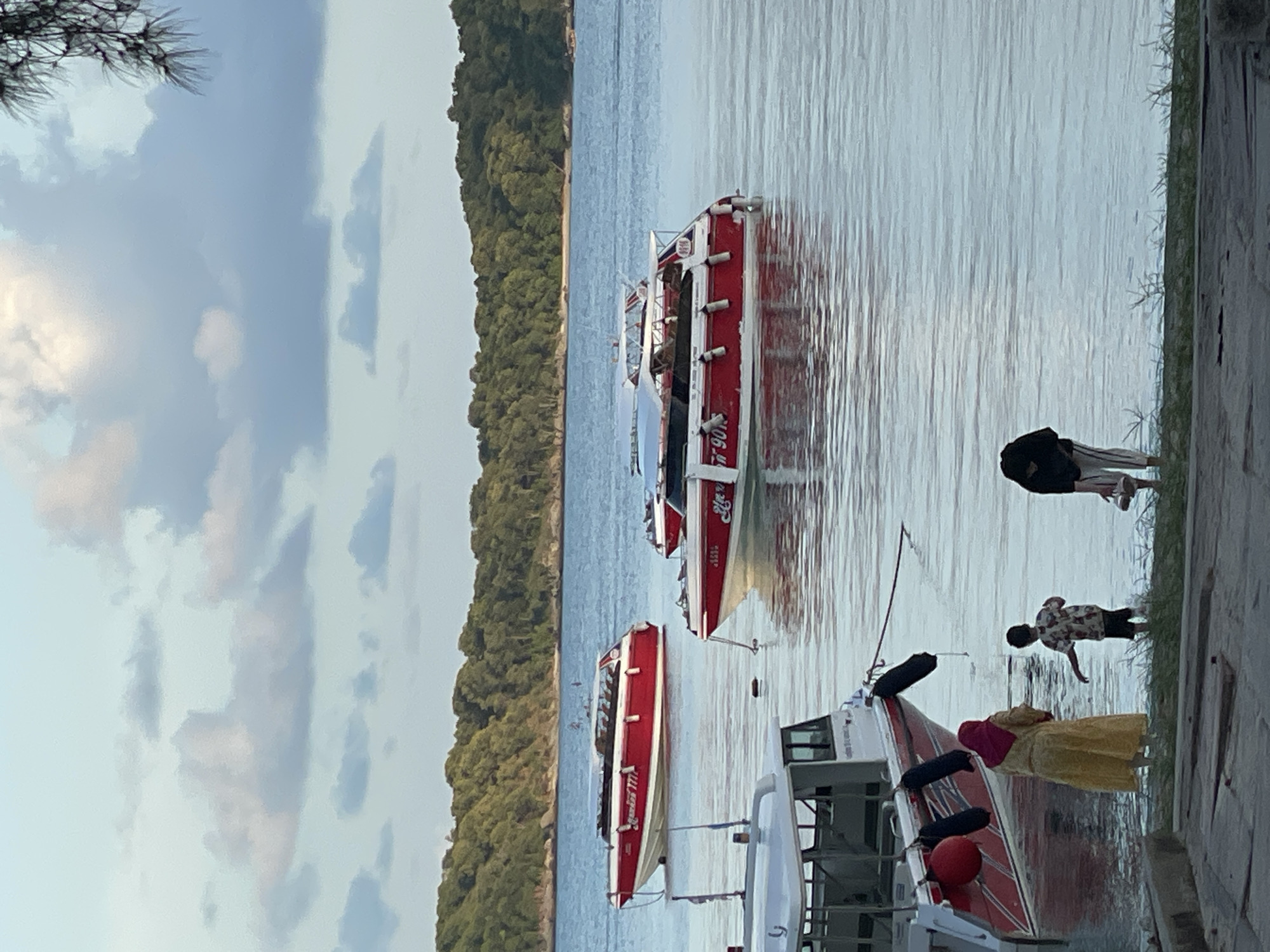 In Rawai, Thailand, on a calm body of water, three red and white speedboats are moored near a treelined shore. The largest speedboat, with a white cabin and red hull, has "EQUISS 777" written on its side. Two other speedboats are positioned further out, also with red and white hulls, one bearing "R 901". In the foreground, on a concrete pier, three individuals are near the water's edge. A woman in a yellow dress and headscarf stands facing away from the camera, looking towards the boats. Beside her, a young boy in a patterned shirt and dark shorts stands facing the water, with his right arm extended. To the right of the boy, a person wearing a black top and striped pants bends over, holding a pair of sandals. The sky above is a soft blue with scattered, wispy clouds. The overall scene depicts a serene coastal environment.
