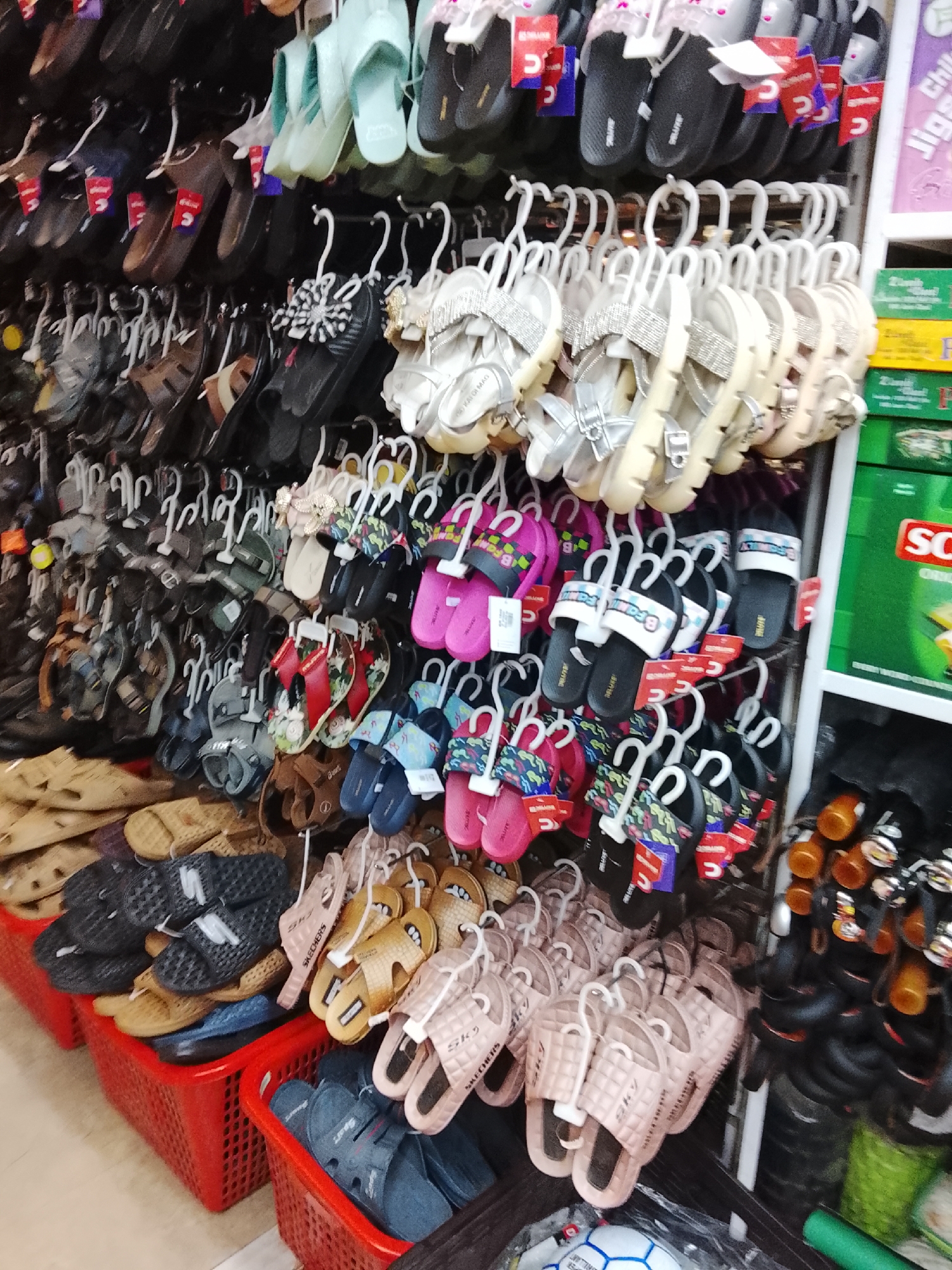 The image shows a variety of sandals and flip-flops displayed for sale at what appears to be a retail store in Gulgasht Colony, Pakistan. The sandals and flip-flops, which come in a wide array of colors and styles, are hung on hooks or placed in red plastic baskets along the aisle. The brand Skechers is visible on some of the footwear. Near the right side, shelves hold other items including drinks. The general environment suggests a typical commercial setting, possibly a department store or a local market in Pakistan.