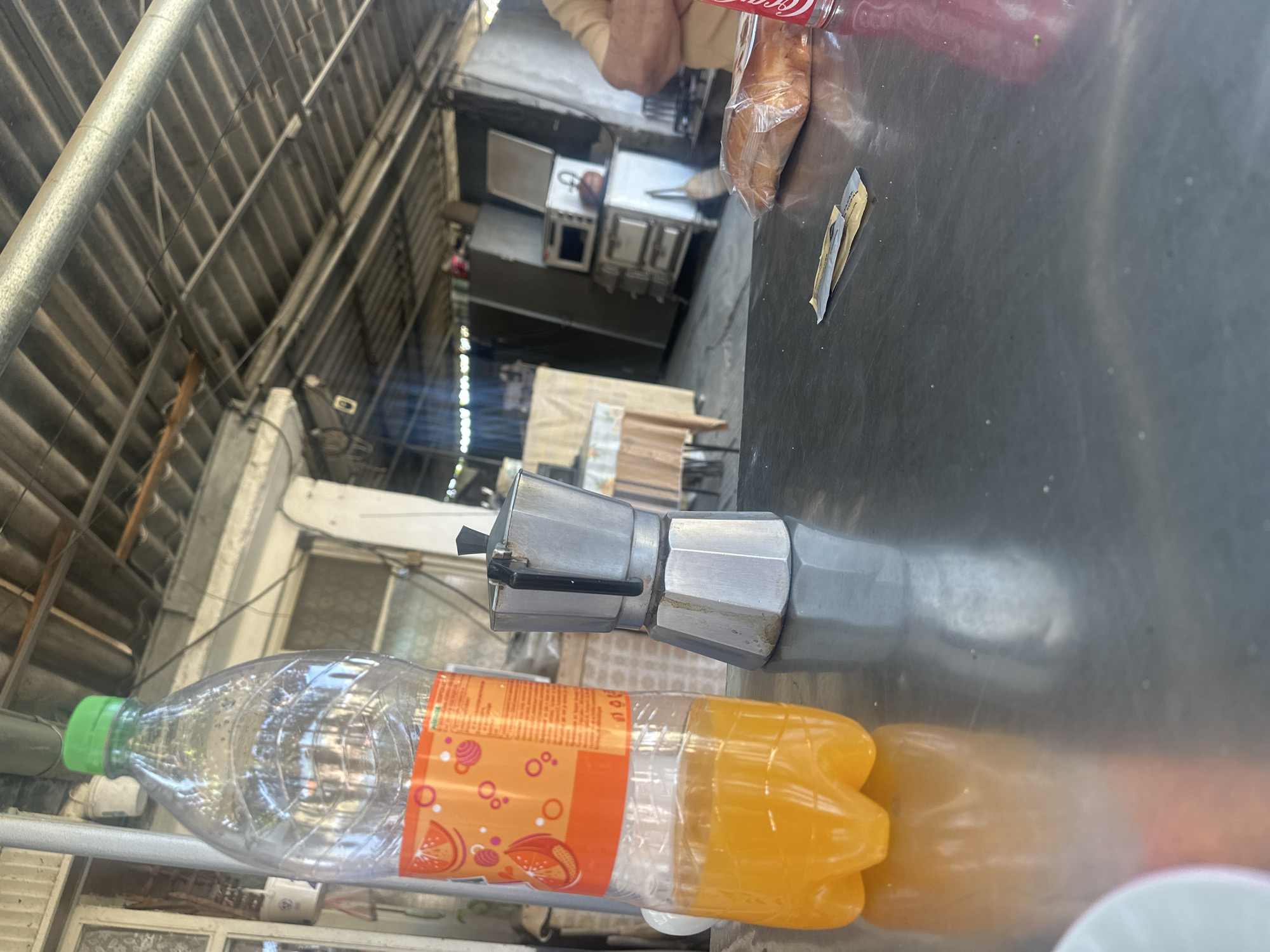 The image depicts a table setting, potentially outdoors, in Razhevo Konare, Bulgaria. On the table are a plastic bottle filled with yellow liquid, a Moka pot, a packaged pastry, and a Coca-Cola bottle. The setting appears casual, possibly a quick meal or snack in an open-air environment. In the background, there's a glimpse of a kitchen or restaurant setting, with metal shelving and cooking equipment.
