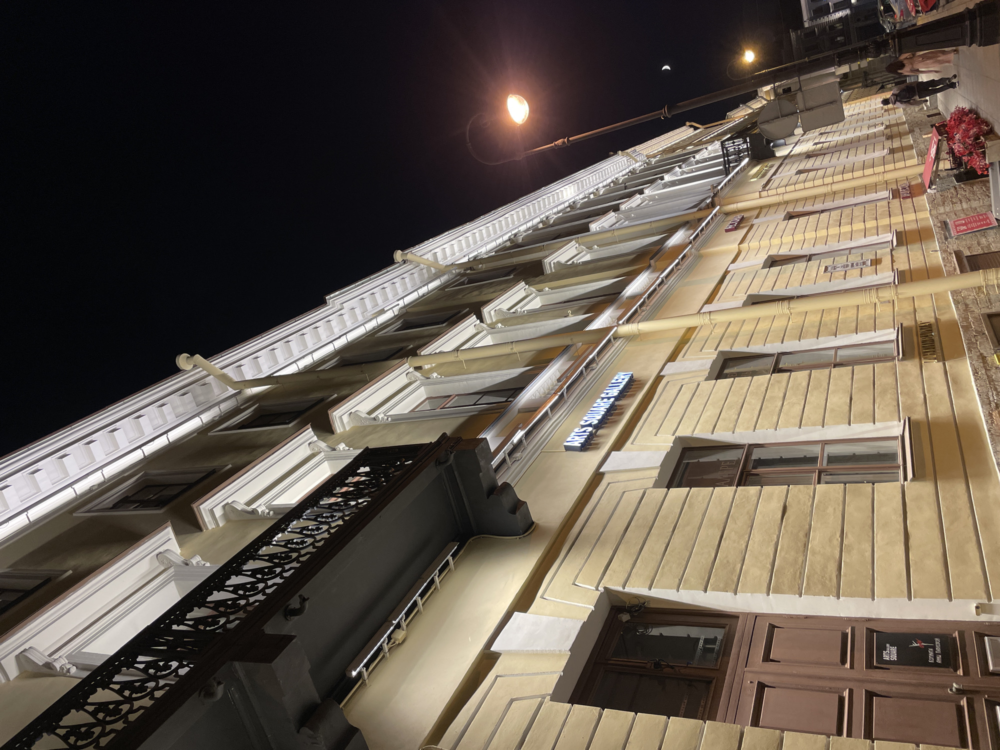 A low-angle, color photograph captures a building at night, situated in Saint Petersburg, Russia. The building's façade, illuminated by artificial light, displays a yellow-toned exterior with white architectural accents. Windows are interspersed along its height. A decorative balcony with a wrought-iron railing projects from the building.
A street lamp, glowing brightly, is visible in the upper portion of the frame, and a crescent moon is just above the top right corner of the building. A sign reading "ARTS SQUARE GALLERY" is attached to the building.
At the top right edge of the image, pedestrians and traffic can be seen in the street. A traffic sign is also visible. The image is taken from a perspective that emphasizes the building's verticality, making the perspective slightly dizzying.