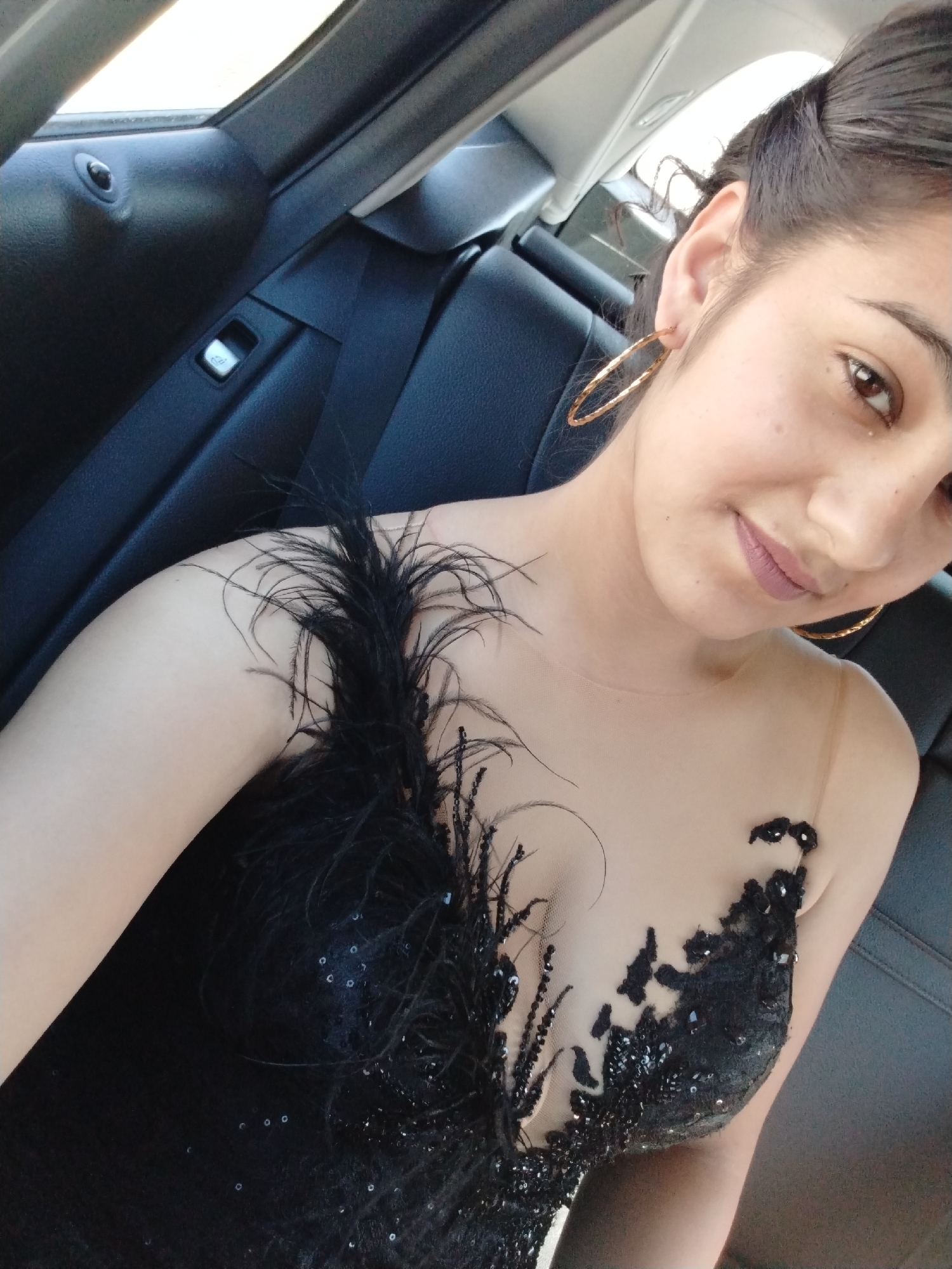 A woman in a black sequined and feathered dress poses for a selfie inside a car. She is wearing large gold hoop earrings and has her hair pulled back. The car interior, with dark leather seats and a seatbelt, is visible in the background. The lighting suggests an outdoor setting during the daytime.
