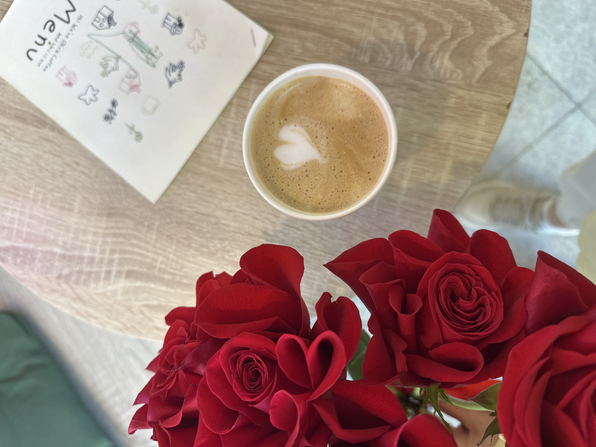 A close-up, high-angle shot shows a cafe scene. Dominating the lower portion are vibrant red roses arranged in a bouquet. A cup of coffee with a latte art heart sits above the flowers. A menu is positioned to the upper left of the coffee, with its corner visible, the word "Menu" readable. The setting is a light wooden table. The lower right corner shows a partial view of a shoe. The overall scene suggests an intimate, aesthetic environment. The location is likely not able to be determined from this photo.