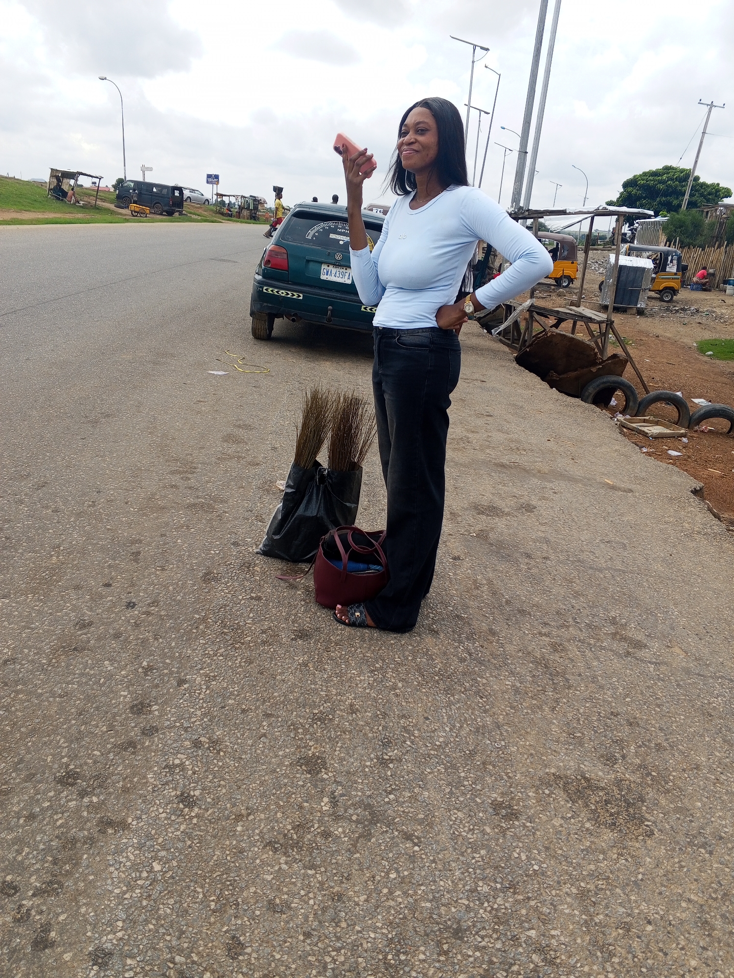 A young Black woman stands on a paved road, holding a pink mobile phone to her ear. She is wearing a light blue, long-sleeved shirt and black pants. To her left are two black plastic bags containing brooms. A dark red purse rests on the ground next to the bags. In the background, a green Volkswagen car is parked. Beyond the car, a makeshift market stalls are visible, with several yellow tricycles, known as 'keke napep', parked nearby. The environment suggests a street market in Pasere, Nigeria, characterized by an overcast sky and a slightly unkempt roadside with debris and discarded tires.