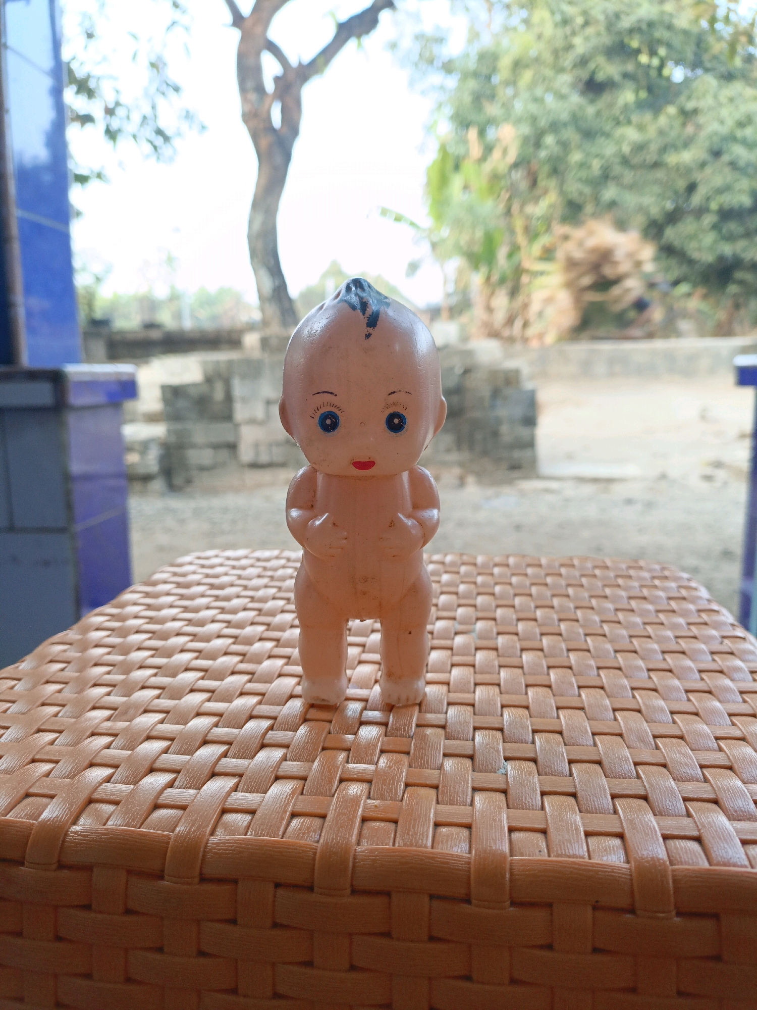 An eye-level shot shows a plastic baby doll standing on a woven orange plastic surface. The doll is light-skinned with blue eyes, a small red mouth, and a tuft of black hair on its head. The background is blurred and depicts an outdoor scene with a large tree, vegetation, and what appear to be cinder blocks, consistent with the environment in Jenu, Indonesia. The lighting suggests it is daytime.