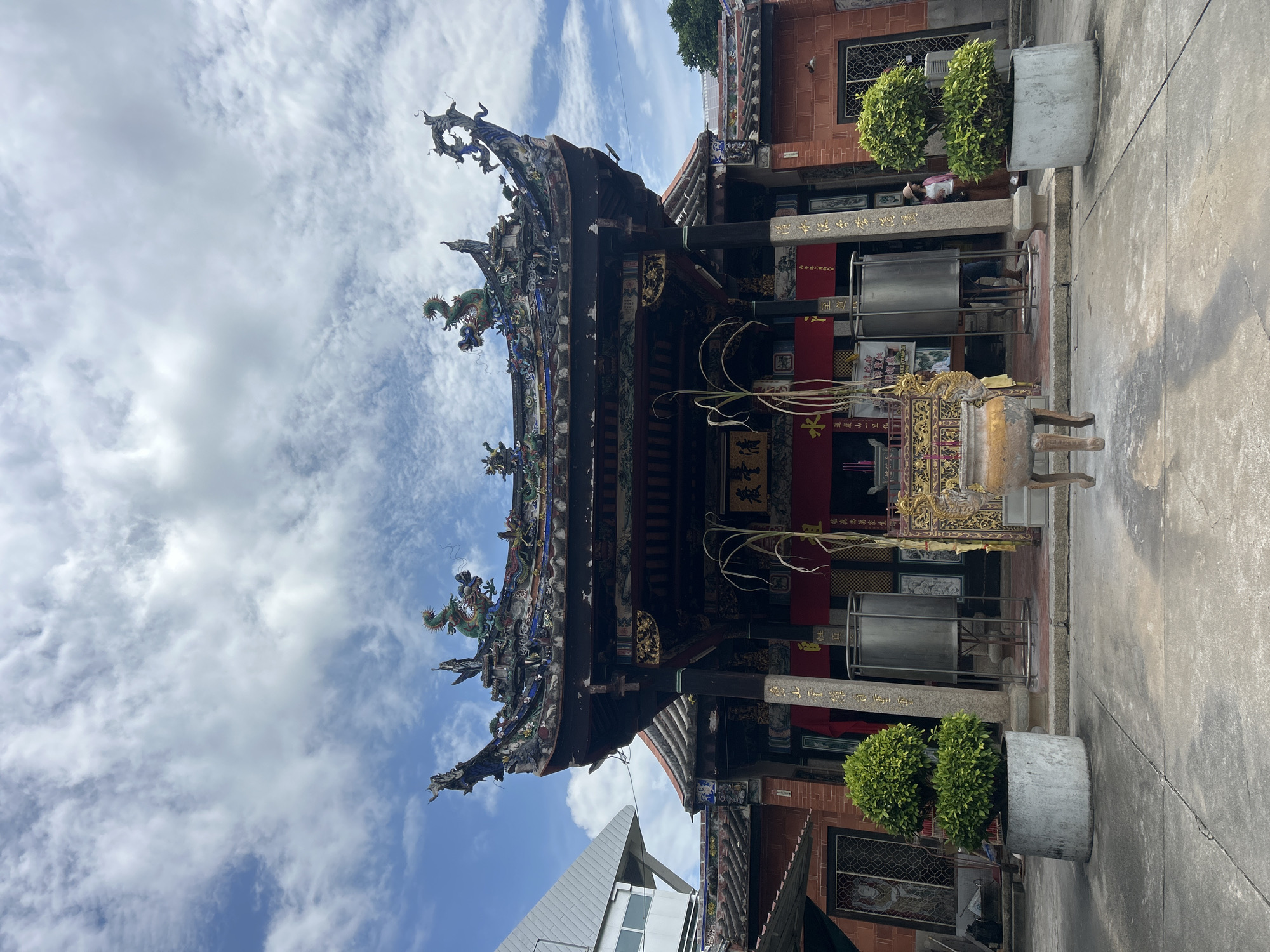 The image shows the exterior of a Chinese temple in George Town, Malaysia, under a partly cloudy sky. The temple features a traditional ornate roof decorated with colorful ceramic figures, including dragons. A large incense burner is positioned in the foreground. Two potted green shrubs flank the entrance, which is flanked by tall pillars. Red curtains hang behind the entrance, obscuring the view inside. A woman in a hat and light-colored top is visible to the right of the entrance, near a potted shrub. The courtyard in front of the temple is paved with concrete. The architecture suggests a historical building, consistent with the many heritage sites found in George Town.