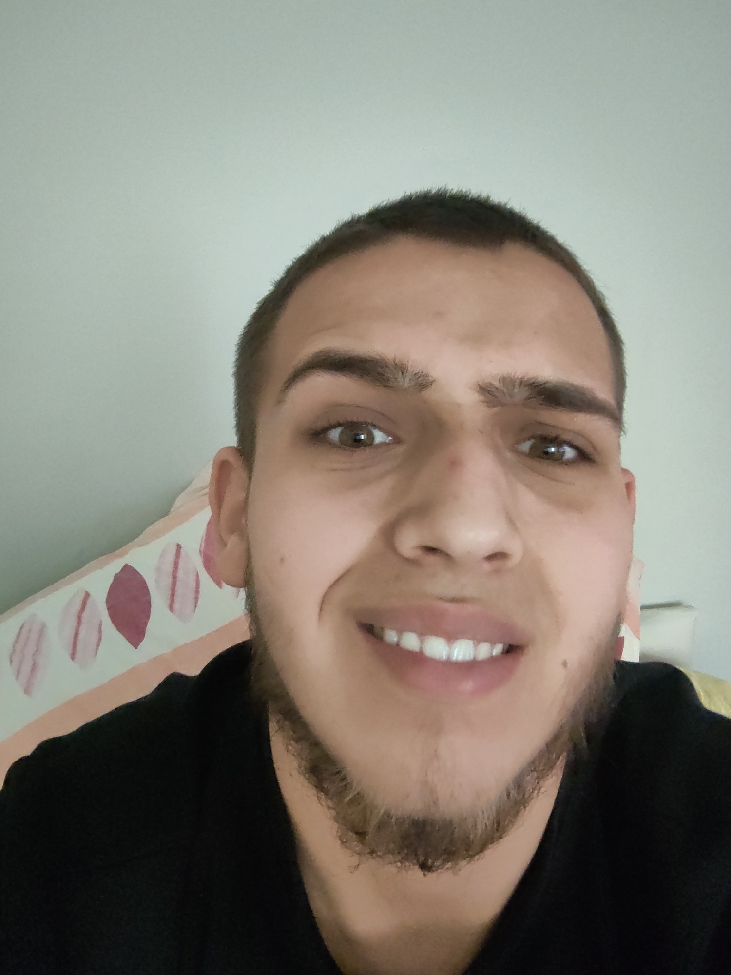 A close-up selfie of a young man. He has short, dark brown hair and brown eyes. He is smiling, revealing white teeth. He has a patchy beard and light skin. He is wearing a black shirt. Behind him is a light-colored wall and a pillow with a leaf pattern. There is no information to determine the city of Sofia, Bulgaria.