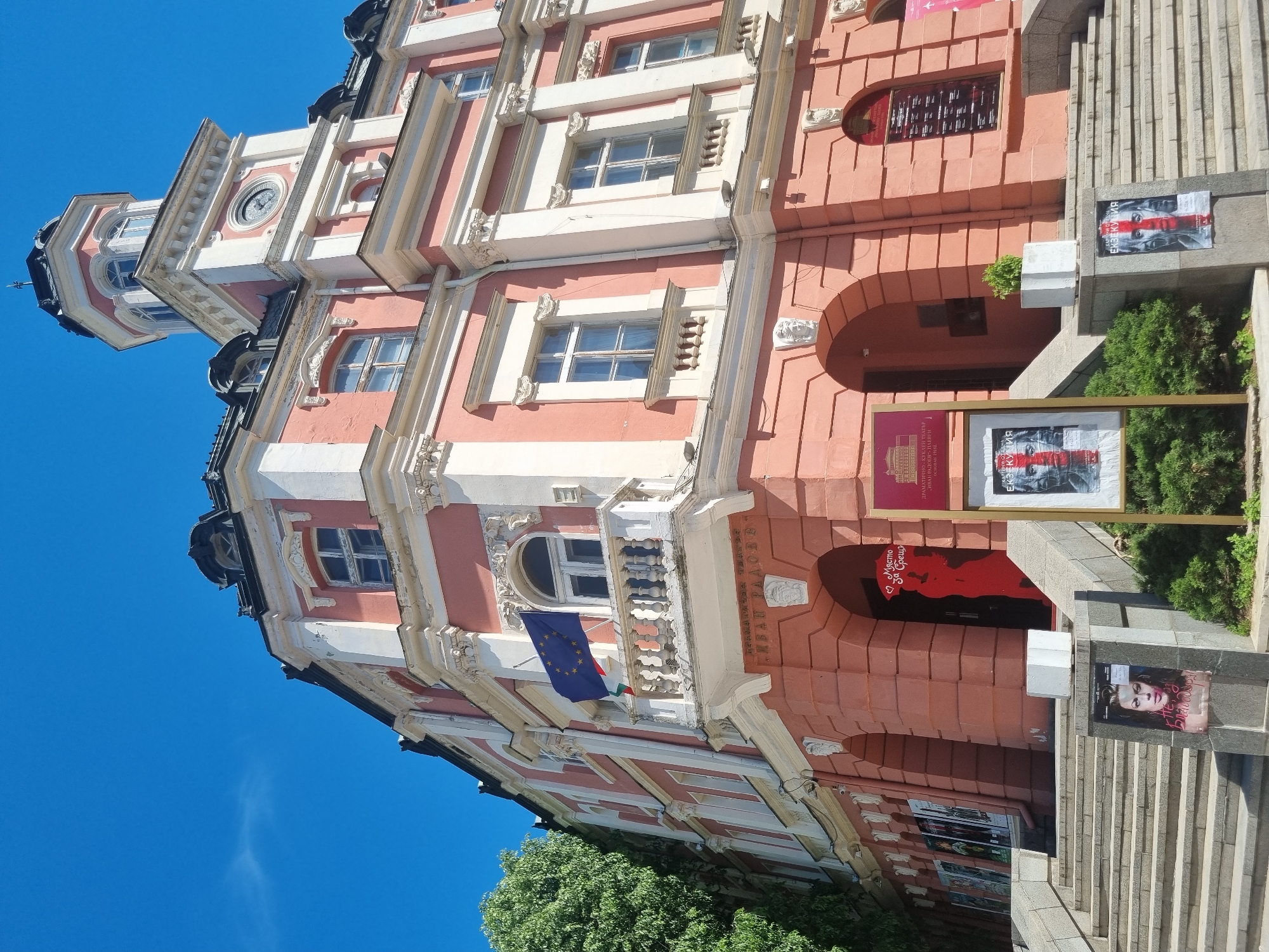 The image depicts the facade of the "Ivan Radoev" Drama and Puppet Theatre in Pleven, Bulgaria. The building is a multi-story structure with pink and white ornate architectural details. There are windows on each level, some of which are arched. An EU flag is visible on one of the balconies. A sign for the theatre is prominently displayed next to the entrance, as well as posters advertising a performance called "Ekzekutsiya" (Execution). The theater's name, "Ivan Radoev," is inscribed on the building. A set of stairs leads up to the entrance. There is greenery and trees at the base of the building. The sky is blue and clear.