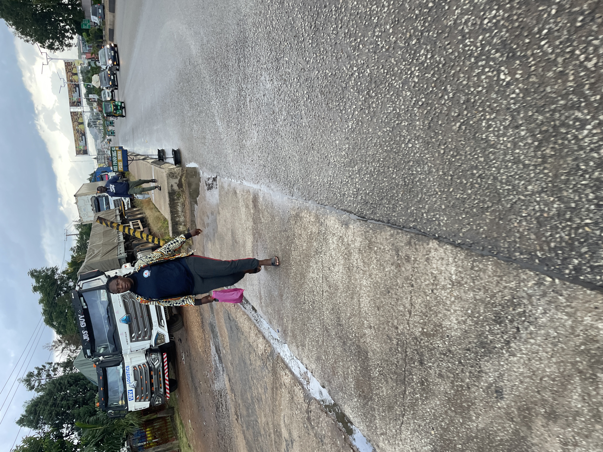 The image shows a street scene in Jos, Nigeria. In the foreground, a woman is walking on a concrete path alongside a wet road. She is wearing dark pants with a red stripe and a colorful jacket over a dark shirt, and carrying a pink bag. Behind her, several large trucks are parked. One truck has "UBV LOGISTICS" written on it, and another has "JOS GUY" on its windshield. In the background, there is traffic on the road, including cars and a tuk-tuk. The sky is overcast. A sign that reads "NO PARKING POLICE ORDER" is visible on the side of the road. The scene suggests a busy urban environment with commercial activity.