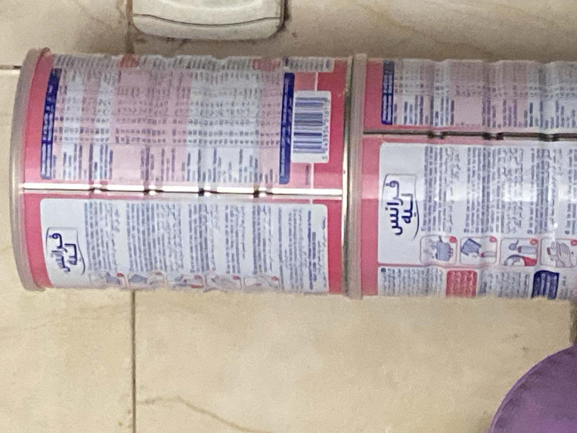 The image shows two stacked cans of baby formula. The cans have pink lids and are white with pink and blue lettering. The upper can has a white label with blue Arabic text that reads "فرانس لنه" and a barcode below it. The lower can also has a white label with blue Arabic text that reads "فرانس لنه". Both cans are positioned vertically against a tiled wall. The wall appears to be a light tan or beige color and has horizontal grout lines. To the right of the cans, a portion of a white appliance, possibly a dispenser or housing, is visible. In the lower left corner of the image, a blurred section of a purple object, likely fabric, is present. The scene is indoors, with the lighting suggesting it's daytime. The overall impression is one of a domestic or commercial storage area. The image is taken from a slightly upward angle, looking at the cans. The environment appears to be in Khan Yunis, Palestinian Territories, as indicated by the context.