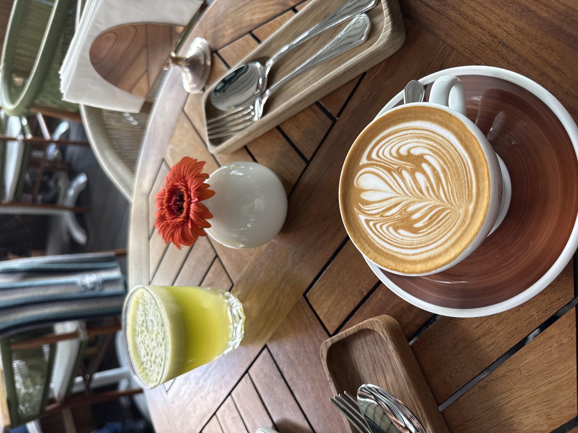 The image shows a wooden table setting at a cafe or restaurant. A cup of latte with latte art sits on a brown saucer, accompanied by a small silver spoon. A clear glass containing a yellow liquid, possibly juice, is also present. Nearby, a white vase holds an orange flower. Cutlery (fork and spoons) are arranged in small wooden trays. A napkin holder with napkins is visible. The location is not discernible from the image, and the claim of it being in Khimki, Russia, cannot be substantiated.
