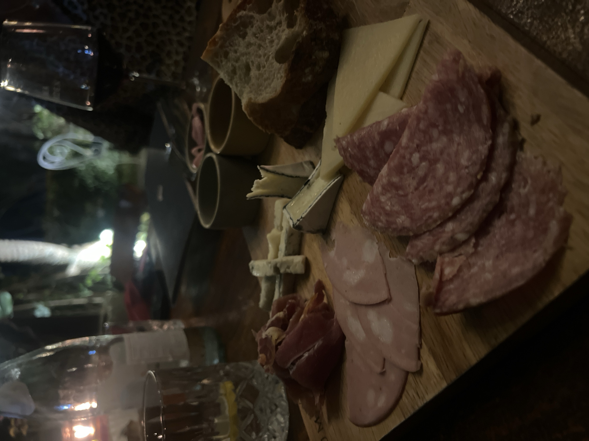 The image depicts a close-up of a charcuterie board at a dimly lit restaurant, likely in North Kuta, Indonesia. The board features an assortment of cured meats, including thinly sliced salami and mortadella, as well as prosciutto. Several types of cheese are present, including a crumbly blue cheese and a wedge of brie-style cheese. Slices of bread are also visible. In the background, a glass of red wine, a bottle of water with a lemon slice, and small bowls containing dips are visible. The overall ambiance suggests a relaxed dining experience.