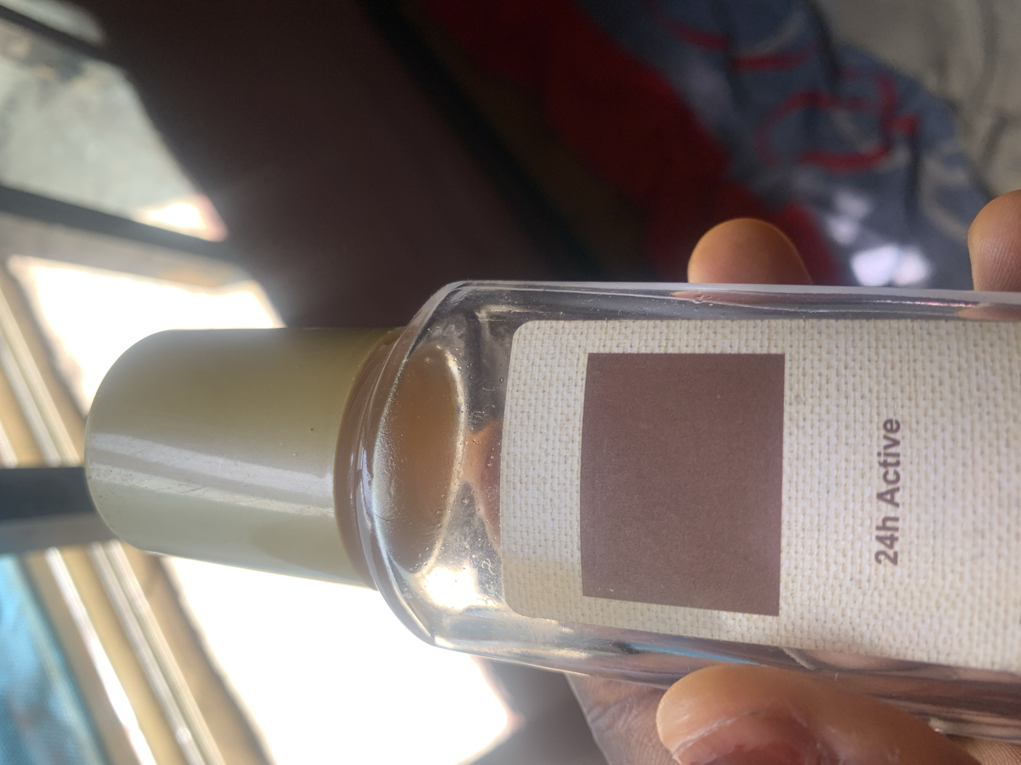 A close-up, low-angle shot shows a person's hand holding a clear glass bottle with a gold cap. The bottle has a beige label with a darker rectangular patch and text that reads "24h Active." The background is out of focus and appears to be indoors, with sunlight streaming through a window. There are no discernible features that would allow for localization to Jauro Yinu, Nigeria.