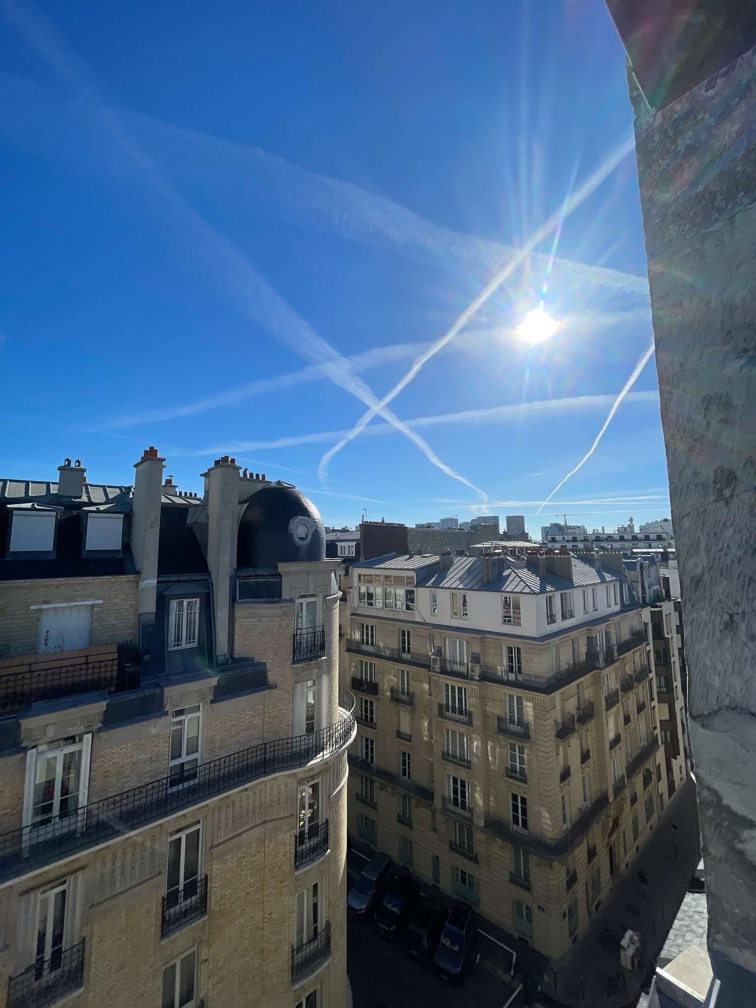 This is a high-angle, wide shot of a Parisian cityscape under a bright, clear blue sky. The sun is high and directly visible, emitting prominent lens flares and rays, indicating a sunny day. Numerous contrails from aircraft crisscross the sky, forming an X-like pattern near the sun.
In the foreground, on the left, is the top of a Parisian Haussmannian-style building, characterized by its mansard roof, dormer windows with shutters, and numerous chimneys. The facade is a pale yellow brick with stone accents. Adjacent to it, and slightly further back, is another building with a darker, domed roof structure and similar architectural features, including dormer windows and chimneys.
The midground showcases a series of apartment buildings, typical of Parisian architecture, with repeating window patterns, balconies, and tiled roofs. Some of these buildings are lighter in color, while others are a warmer, beige stone. The street level is visible in the lower portion of the frame, where parked cars can be seen.
The right side of the image is dominated by a close-up view of a rough, textured stone surface, possibly part of a building or monument, which partially frames the scene and creates a strong contrast with the smooth lines of the cityscape. The overall impression is one of a sunny day in a densely built urban environment.