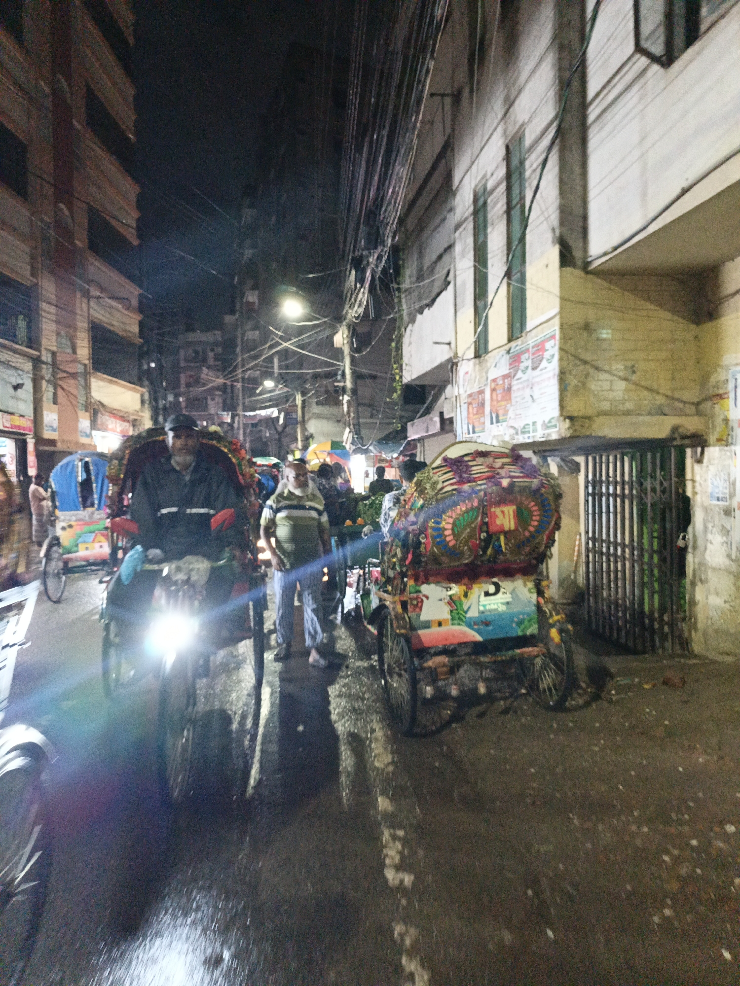 A wet street scene at night in Unknown, Unknown shows several rickshaws with their drivers and other pedestrians. The street is lined with multi-story buildings, some with boarded-up windows or closed metal gates. Electrical wires crisscross overhead. A rickshaw in the foreground has its headlight on, creating a glare. The rickshaws have decorative designs. A man in a striped shirt and light pants stands on the sidewalk.