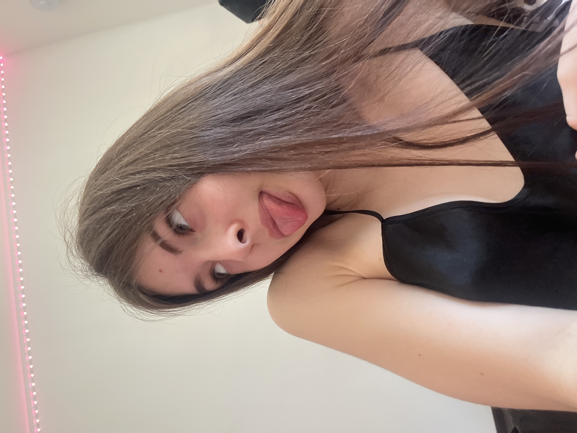 A young woman with long, brown hair is sticking her tongue out, wearing a black tank top, in an indoor setting. A pink LED strip light is visible along a wall. There is no information that indicates the location of the photo is in Saint Petersburg, Russia.