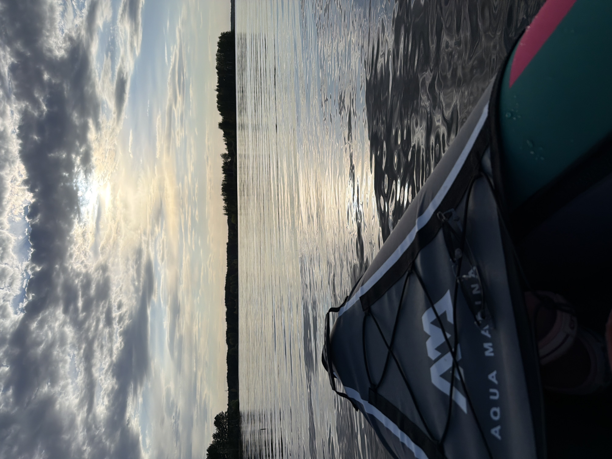 The image depicts a water landscape with a kayak in the foreground. The water reflects the sky, which is filled with clouds. In the distance, there's a line of trees. Part of the kayak is visible on the right side, showing the brand name "AQUA MARINA". The location appears to be a lake or river, possibly near a forested area, given the line of trees in the distance. While it is mentioned that the location is in the city of Chaykovsky, Russia, there is no visual information to confirm this claim from the image. No people or subjects can be identified.