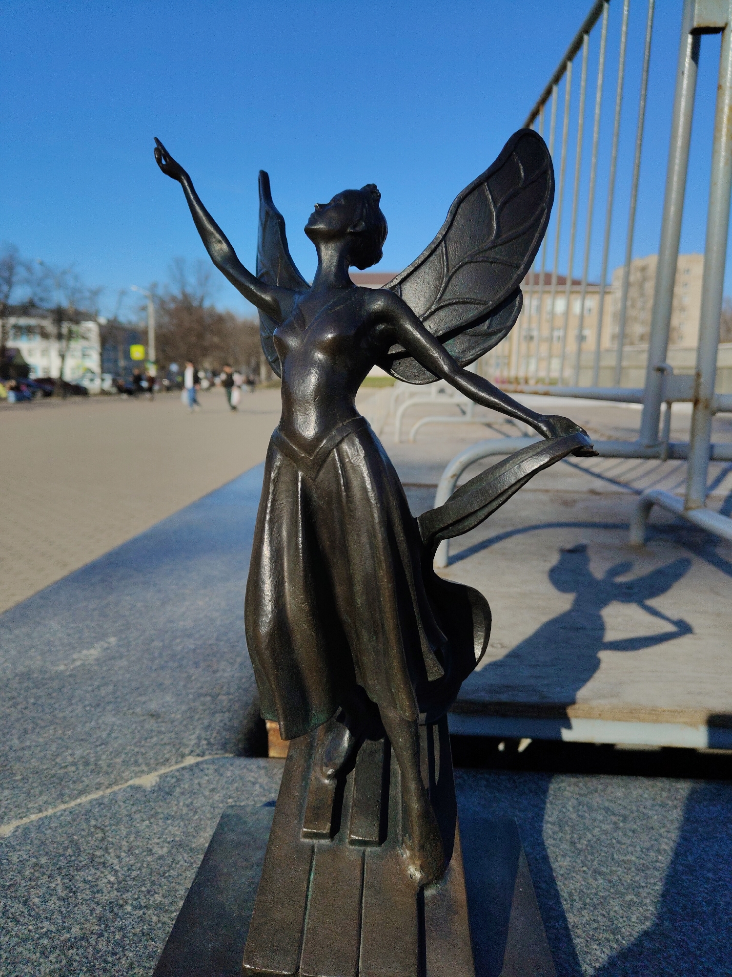 A bronze statue of a winged female figure stands prominently in an outdoor setting under a clear blue sky. The statue, depicting a slender figure with a flowing dress and large, detailed wings, is positioned dynamically, with her head tilted upwards and her right arm extended towards the sky. Her left arm is slightly bent, holding a flowing element that integrates with the base. The base of the statue appears to be designed to resemble piano keys, suggesting a theme related to music or performance.
The statue casts a distinct, dark shadow on the granite-like ground to its right, indicating bright sunlight and a clear day. In the background, a paved pedestrian area stretches into the distance, where several blurred figures of people can be seen walking. To the right of the statue, a tall, grey metal fence or barrier runs vertically, creating a visual backdrop. Further in the background, out-of-focus buildings and bare trees are visible, typical of an urban or park environment. The overall impression is one of aspiration and artistry set against a bright, active public space. There is no visible text.