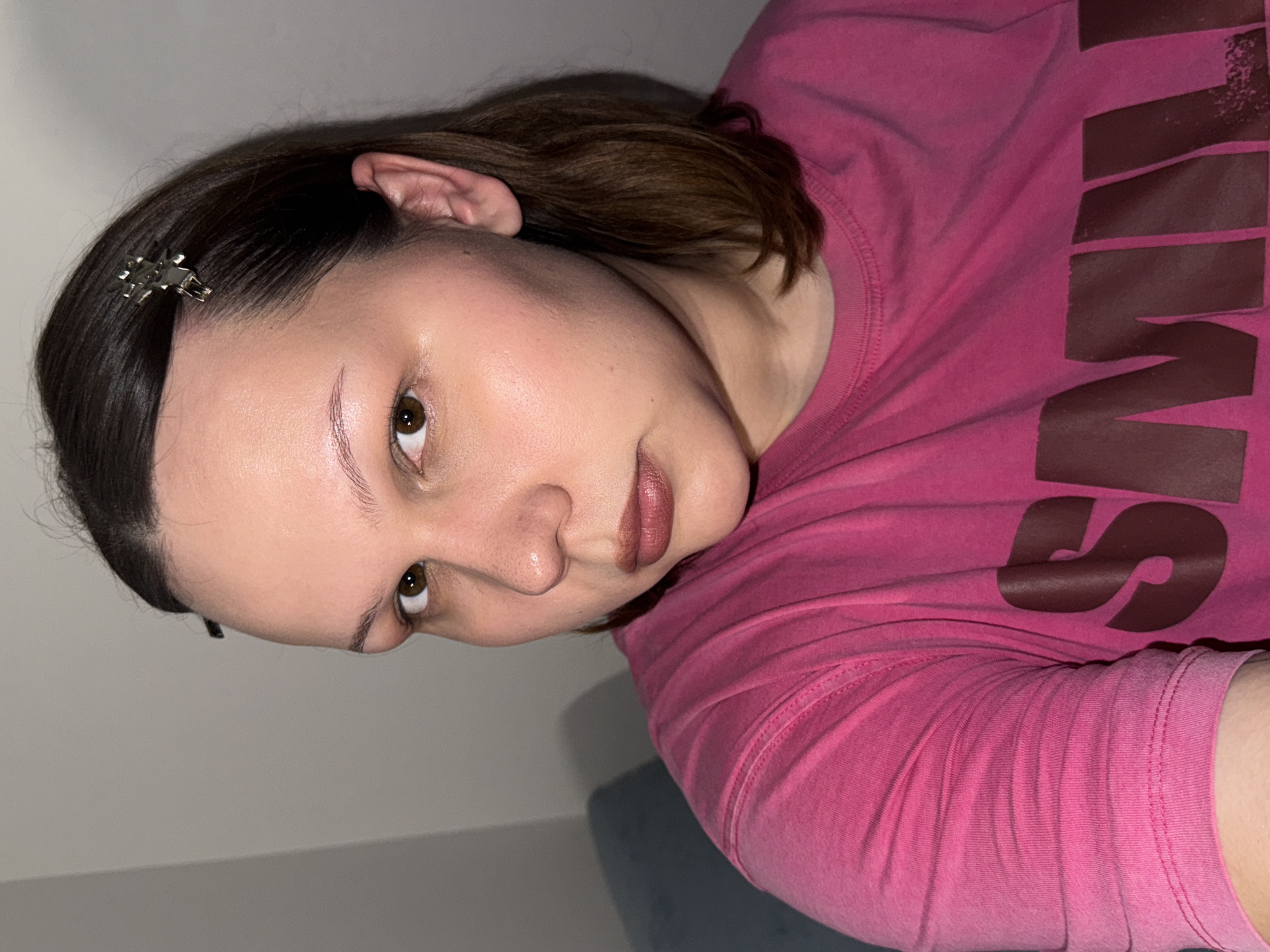 A person wearing a pink shirt is the primary subject of the image. The person has brown hair, and a hair clip is visible in their hair. The background appears to be a wall, and there may be a portion of a piece of furniture visible. The shirt has large dark text on it, but only parts of the letters are visible. The image does not contain information to determine the location is Chaikovsky, Russia.