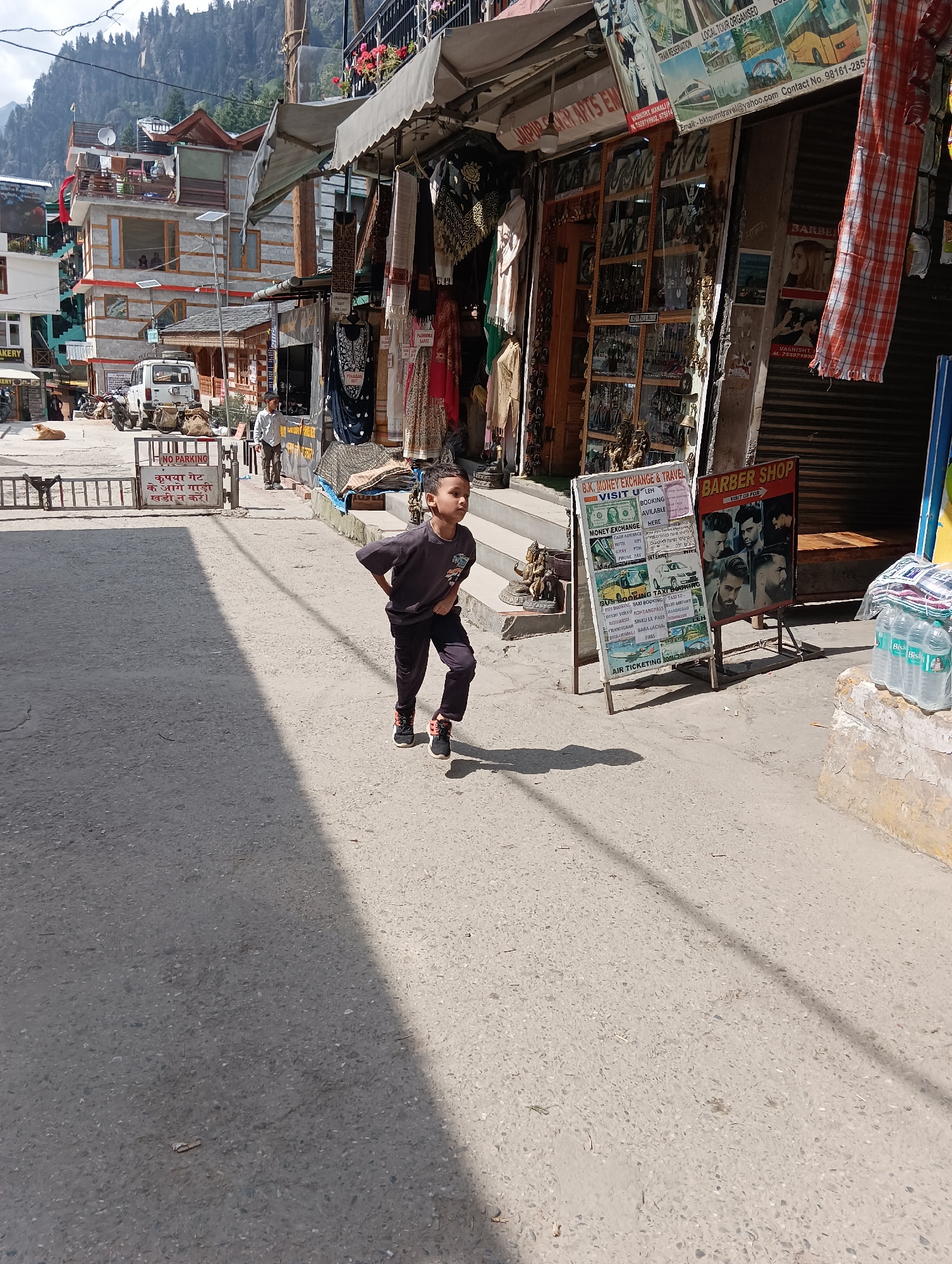 A young boy runs down a paved street in a mountainous town. He is wearing a dark gray t-shirt and dark gray pants. He has his hands on his hips and is looking to the left. The street is lined with shops and buildings. One shop has a sign that says "BARBER SHOP". Another shop has a sign that says "B.K. MONEY EXCHANGE & TRAVEL". There is a white SUV parked on the street and a "NO PARKING" sign. In the background, there are tall, forested mountains. The sun is shining and casting shadows on the street. There is a dog lying down in front of a shop on the left side of the street. A man in a white shirt and light brown pants is walking away from the camera near a parked motorcycle. Another man in a lighter colored shirt and darker pants is visible walking in the distance. A rack of colorful fabrics hangs outside of a shop on the right side of the image. Bottles of water are stacked on a stone ledge in front of the shop.