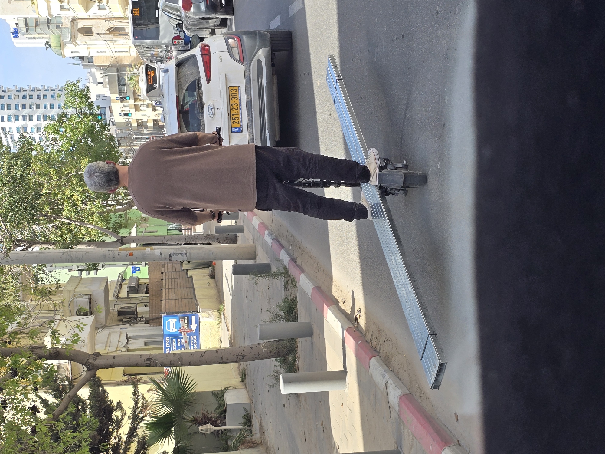 The image shows a man riding an electric scooter on a street in Tel Aviv, Israel. He is wearing a brown shirt and black pants. The scooter is carrying long metal beams. A white car with the license plate number 251:23-303 is parked nearby. A parking sign is visible in the background. The image shows urban infrastructure like buildings, street trees, and parked cars.