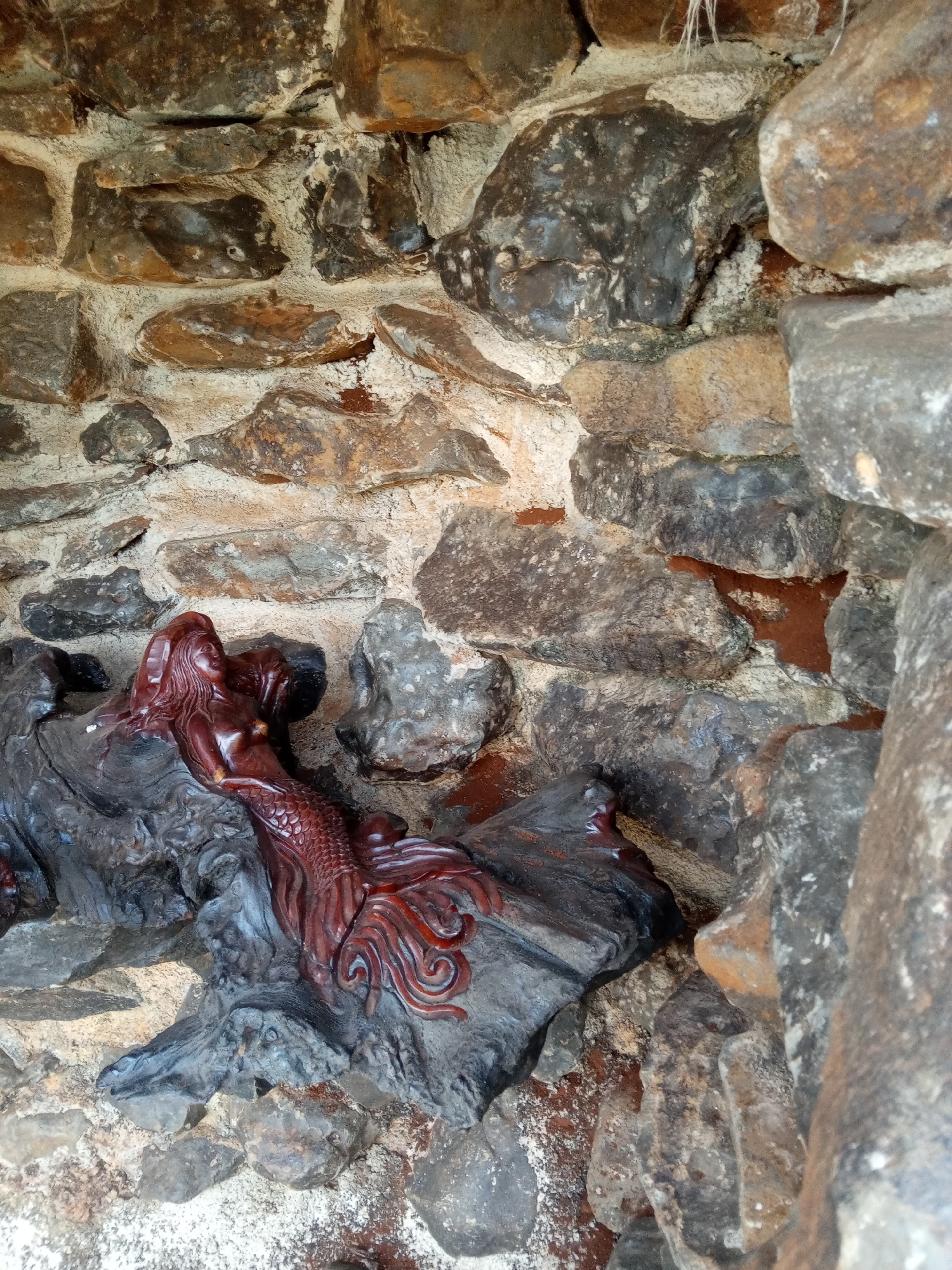 A close-up, eye-level shot depicts a maroon-colored, carved wooden mermaid figure resting on a gray-colored, root-like platform, which is set within a rough stone enclosure or wall. The wall is constructed of varied shapes and sizes of rocks, mostly in shades of gray, brown, and tan, cemented together with a light-colored mortar. The mermaid, positioned on its side, displays intricate details of its anatomy and flowing tail, while the platform beneath has a rough, organic texture. The photograph was taken in the city of Chư Sê, Vietnam.