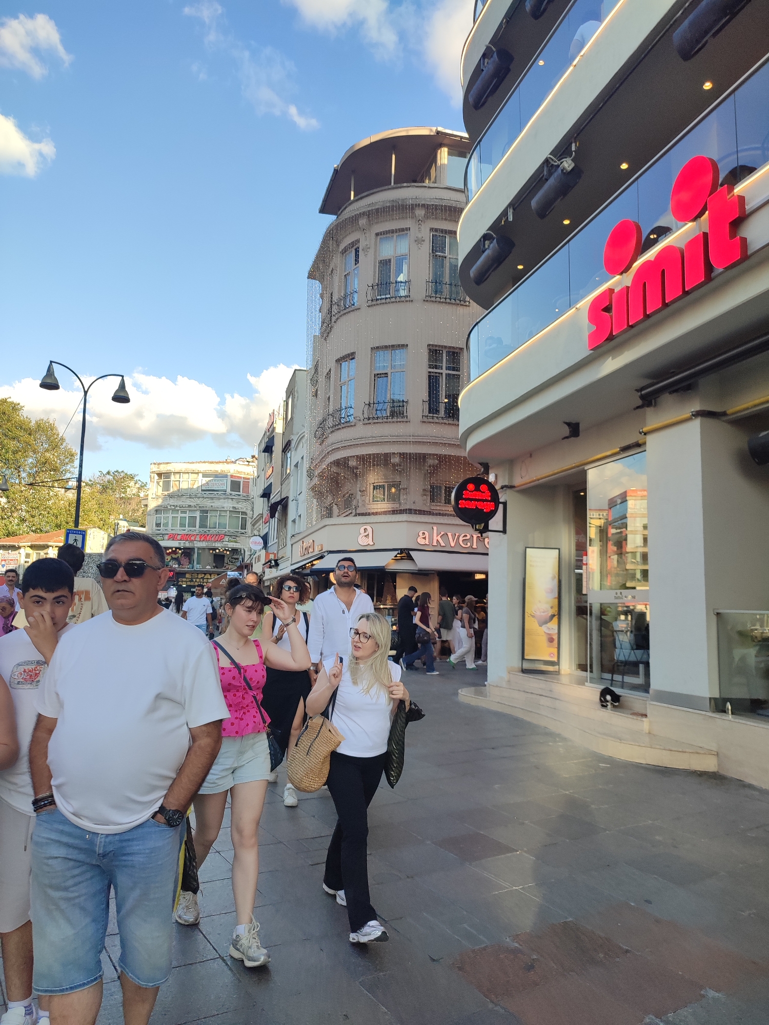 The image depicts a street scene in Kadıköy, Turkey, on a bright, partially cloudy day. The foreground is dominated by a group of people walking on a paved sidewalk. A man in a white t-shirt and blue denim shorts stands prominently on the left, with his hand in his pocket. To his right, a young woman in a pink polka-dot top and light blue shorts walks with a straw bag slung over her shoulder. Further to the right, a woman in a white t-shirt and black pants walks with her hands clasped.
Behind the people, commercial buildings line the street. A modern building with a curved glass facade and red signage reading "simit" is visible on the right. To its left, an older building with arched windows and decorative elements stands. Several streetlights with a distinctive curved design are also present. A sign with a red circular emblem and text that appears to be "simit sarayı" is mounted on a pole. Reflections of buildings and the sky are visible in the glass windows. A black and white cat is present on the steps of the building on the right. The ground is paved with dark stones, with some lighter-colored sections. The overall atmosphere suggests a bustling urban environment.