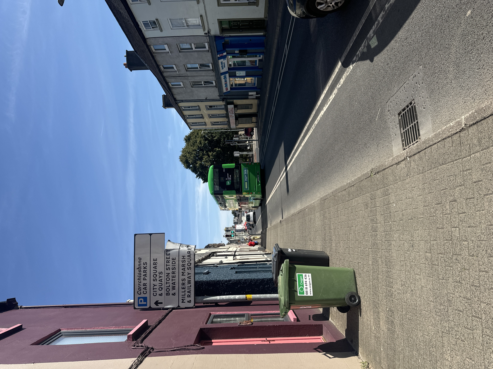 The image shows a street scene in Waterford, Ireland, on a sunny day. The foreground features a paved sidewalk with two large trash bins, one green and one black. The green bin has the "Oxigen" logo and a phone number. A purple building is on the left, with a window and trim visible. A street signpost is located between the building and the sidewalk, indicating directions to "Carrchlósanna CAR PARKS", "CITY SQUARE & QUAYS", "BOLTON STREET & WATERSIDE", and "MILLERS MARSH & RAILWAY SQUARE". A green double-decker bus is in the street, moving away from the camera, with "TFI" and "114 113" visible on its side. Several cars are also on the road, and traffic lights are visible in the distance. The buildings lining the street are typical of Irish town architecture, with shops and apartments. The sky is clear blue with a few faint clouds.