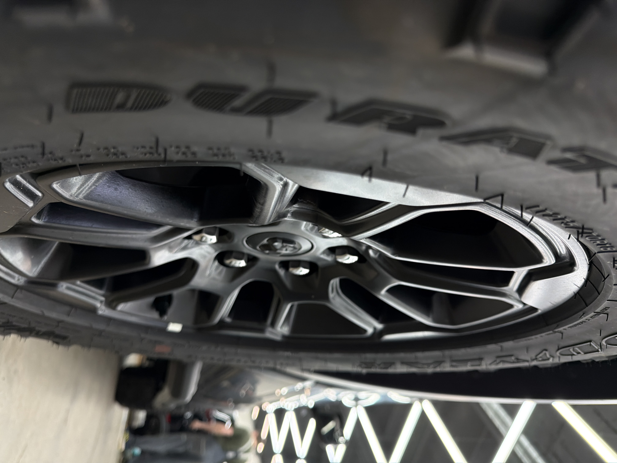 The image displays a close-up, low-angle view of a dark grey or black multi-spoke car wheel and an all-terrain tire, set indoors, likely within a professional garage, showroom, or service center in Krasnoyarsk, Russia. The wheel rim features a modern, angular spoke design, with six metallic lug nuts visible around a central cap. The tire exhibits a robust, chunky tread pattern on its sidewall, indicative of an off-road or all-terrain application.
Visible text on the tire sidewall clearly shows parts of the brand name "GOODYEAR" and the model name "WRANGLER". Other indistinct markings, possibly numerical or technical specifications such as "LT", "285", "33%", and individual letters/numbers like "PP" and "4", are also faintly discernible due to blur and perspective.
The background is highly blurred but reveals numerous bright, parallel fluorescent or LED light strips, which reflect on a polished surface below the vehicle. This lighting arrangement suggests a well-lit and clean indoor environment. No specific activity is taking place; the image captures the static appearance of the wheel, potentially during a display, inspection, or after service. No people are clearly visible. The indoor setting means the time of day and weather are not relevant.
