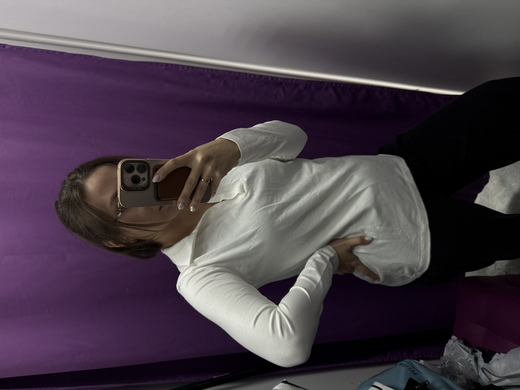 A woman in Pervomaysky, Russia is taking a selfie in a mirror. She is wearing black pants and a white, long-sleeved shirt. Her phone is in a gold case. A purple sheet or backdrop is behind her. There is clutter on the floor including a plastic bag, a blue bag, and a pink box.
