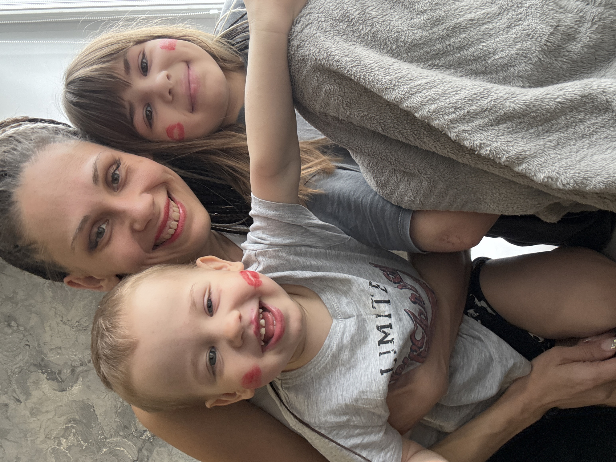 This heartwarming image captures an intimate family moment featuring an adult woman embracing two smiling children, a girl and a boy, in what appears to be an indoor setting during the daytime. The subjects are positioned closely, all looking towards the camera with joyful expressions.
The adult woman, visible on the left, has dark dreadlocks or braids and a bright, toothy smile. The older child, a girl with long, light brown hair and bangs, is nestled above the woman, displaying a sweet, content smile. The younger child, a boy with short, light hair, is in the foreground, below the woman, with a wide, energetic grin that reveals his teeth. Both children playfully have distinct red lipstick kiss marks on their cheeks, suggesting affectionate interaction.
A soft, light grey fluffy blanket or towel is draped over the individuals, contributing to a cozy and warm atmosphere. In the background, soft daylight streams in from a window, and a patterned surface, possibly a wall or floor with a marbled or textured design, is visible. The boy is wearing a light grey t-shirt on which a partial word, "LIMIT," and a graphic design resembling an anchor are discernible. The overall impression is one of happiness, love, and playful family connection.