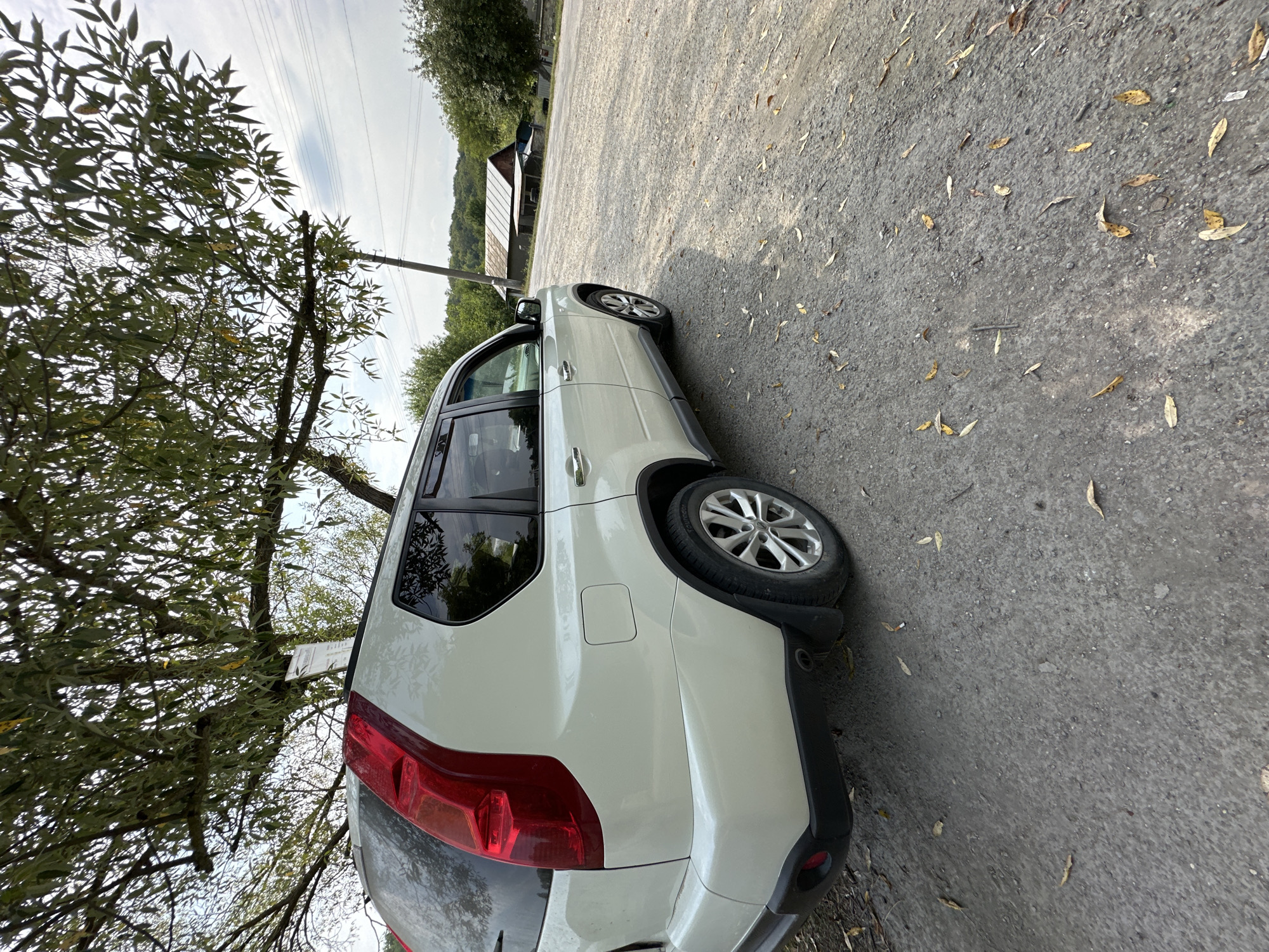 A white 2007-2013 Nissan X-Trail is parked on a gravel road under a large tree. The vehicle is facing away from the camera and slightly to the left, with its rear passenger side in view. The car has a red taillight assembly and a silver alloy wheel. Scattered dry leaves are present on the ground around the vehicle. In the background, there is a rural landscape with green trees, a building with a corrugated metal roof, and power lines. The sky is cloudy. The location is the city of Makiv, Ukraine.