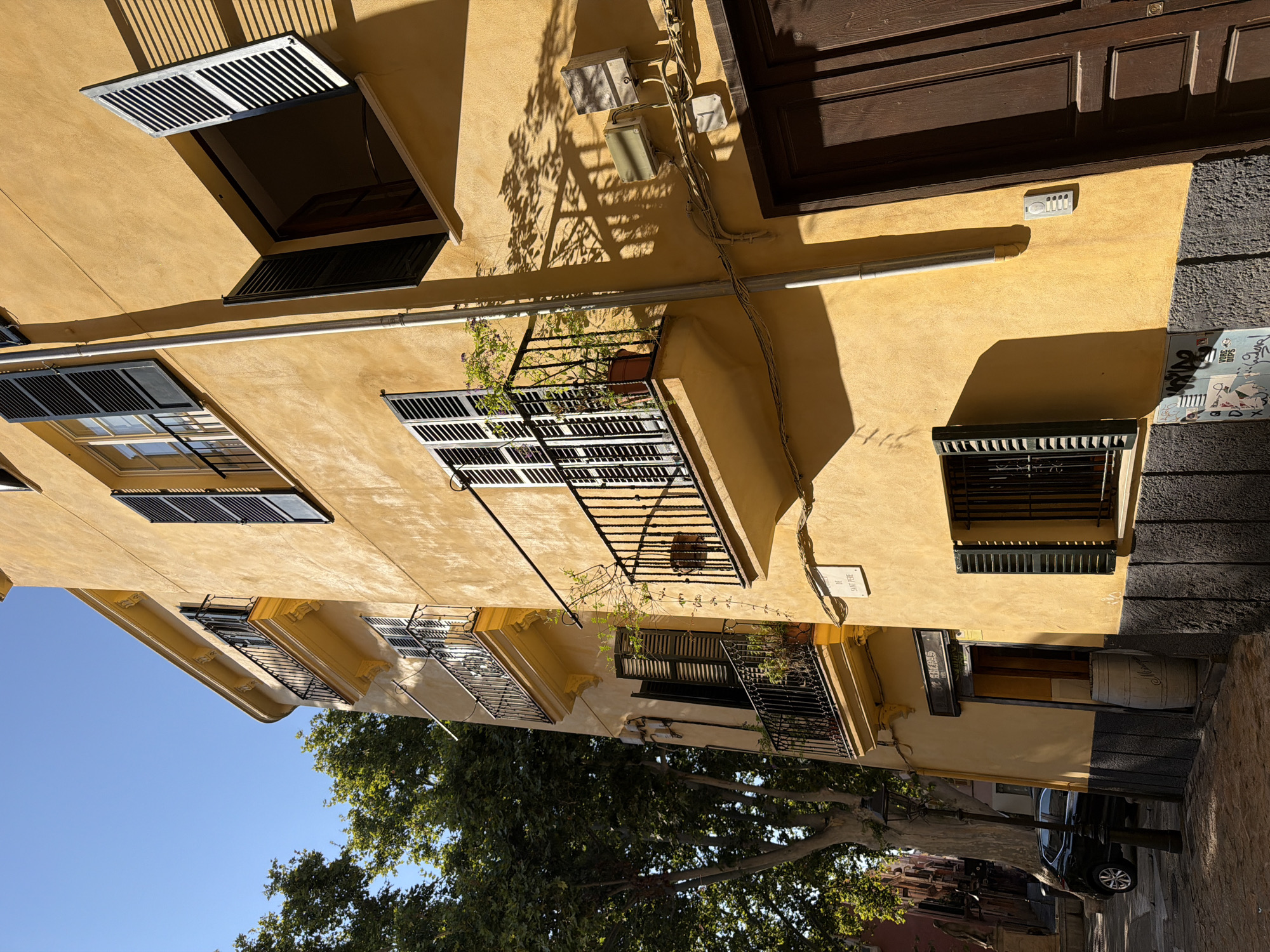 The image shows a sunlit street in Palma de Mallorca, Spain, focusing on a yellow building with several balconies and dark shutters. A black car is parked on the cobblestone street to the left. The building has multiple stories, with black wrought-iron railings on the balconies. One balcony on the upper floor has an open window with shutters and a plant in a pot. Another balcony below it also features a plant. The facade of the building is textured and has some visible electrical conduits and boxes. A wooden door is visible on the right side of the building. The street is paved with cobblestones, and a street lamp stands on the left. There are shadows cast by trees and the building itself. A white sign with black lettering, reading "DE SANT PERE," is attached to the building facade. There are no people visible in the image.