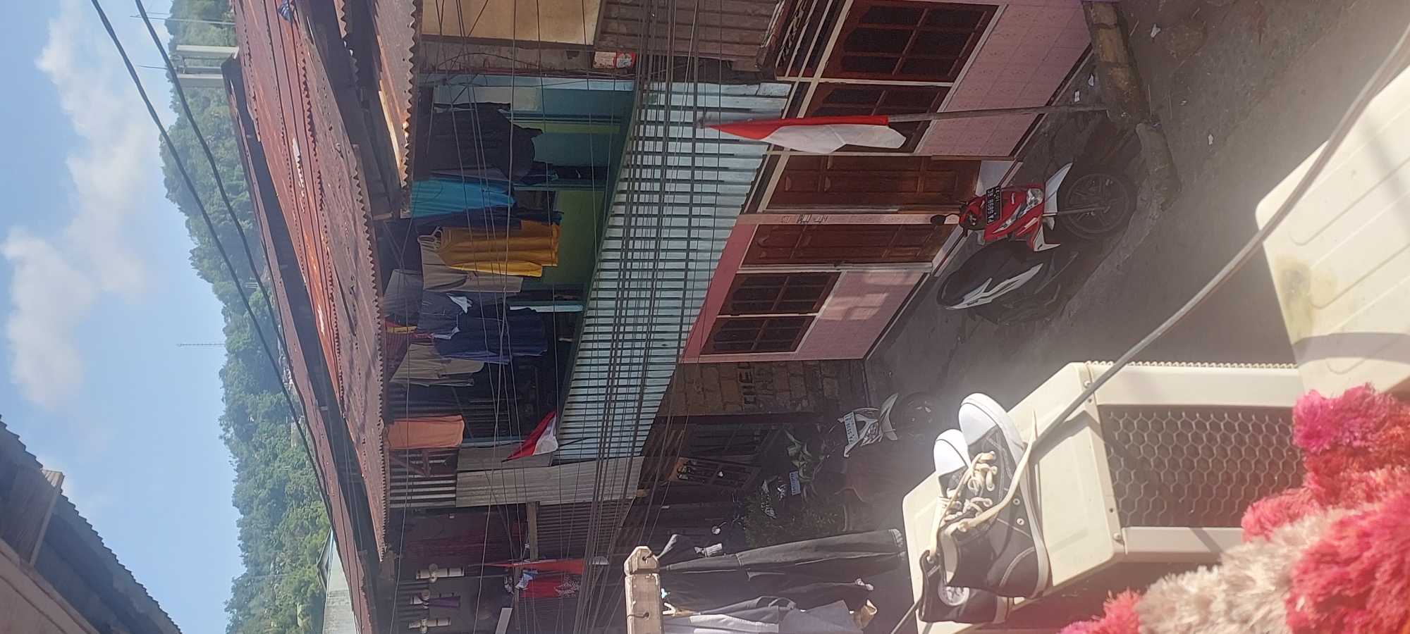 This is a high-angle, full shot of a residential street scene in the city of Jayapura, Indonesia, on a clear, sunny day. The focus is on the multi-story buildings with corrugated tin roofs, typical of the area. Laundry is hung out to dry on lines stretched across the balconies and windows of the upper floors. A red and white Indonesian flag is displayed prominently on a pole attached to one of the buildings.
In the foreground, a pair of high-top sneakers rests on top of an air conditioning unit. The street level shows a couple of parked motorcycles, one red and white, and another white one partially visible. The buildings themselves are painted in various colors, with wooden doors and window frames. The scene conveys a sense of everyday life in a densely populated urban environment, with the natural landscape of a lush green hill forming the backdrop. The overall impression is one of a vibrant, lived-in community.