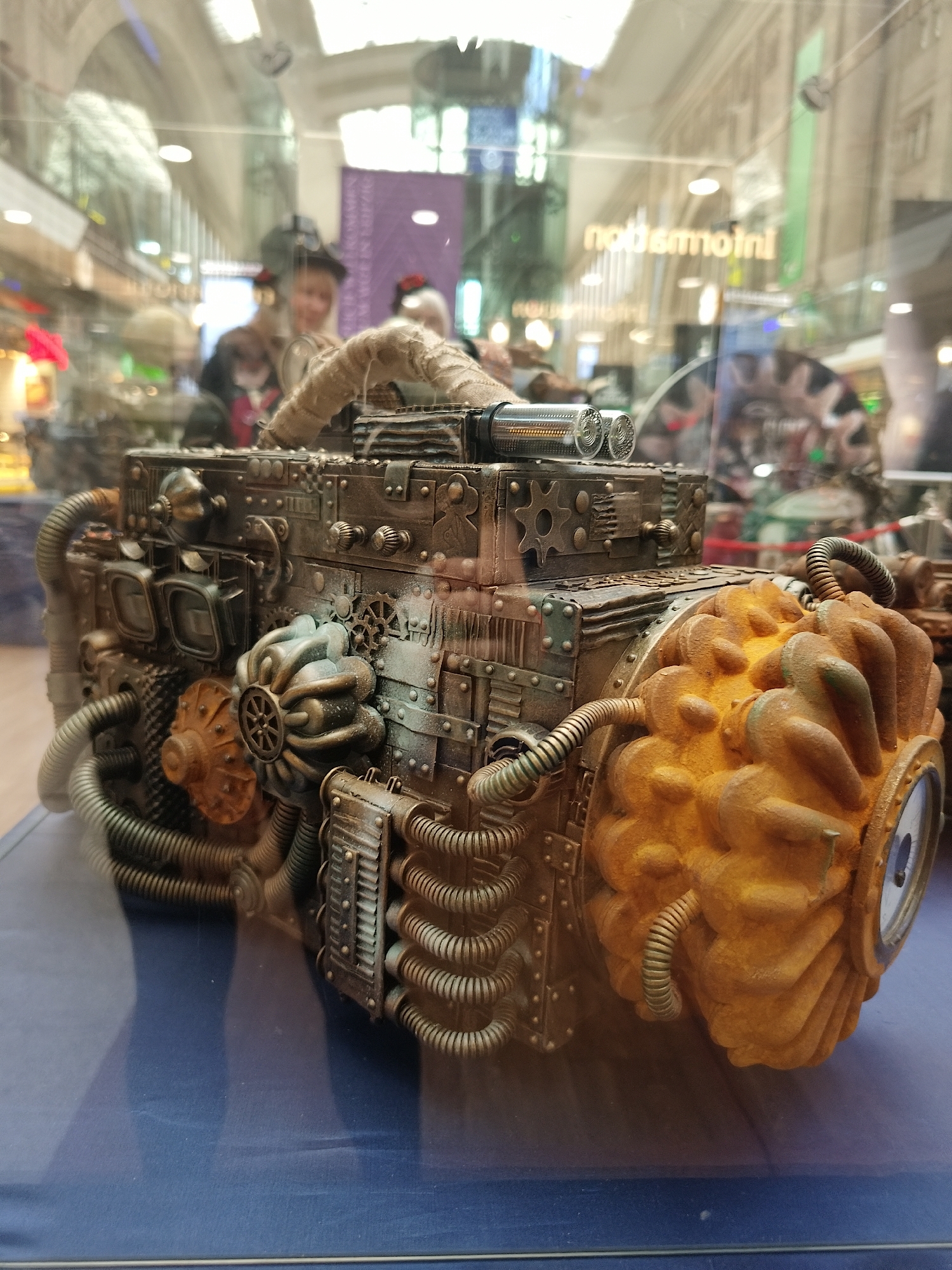 The image shows a steampunk-style decorative object resembling a machine, displayed inside a glass case. The machine is predominantly brown and features various mechanical elements such as gears, pipes, and a large, textured, orange-colored front piece with a round opening. The background shows a shopping center or similar indoor location, with reflections and blurred figures visible through the glass. The location is a store in Leipzig, Germany. The subject of the display case is steampunk-inspired art. There are people shopping in the background. The environment is a brightly lit indoor retail space.