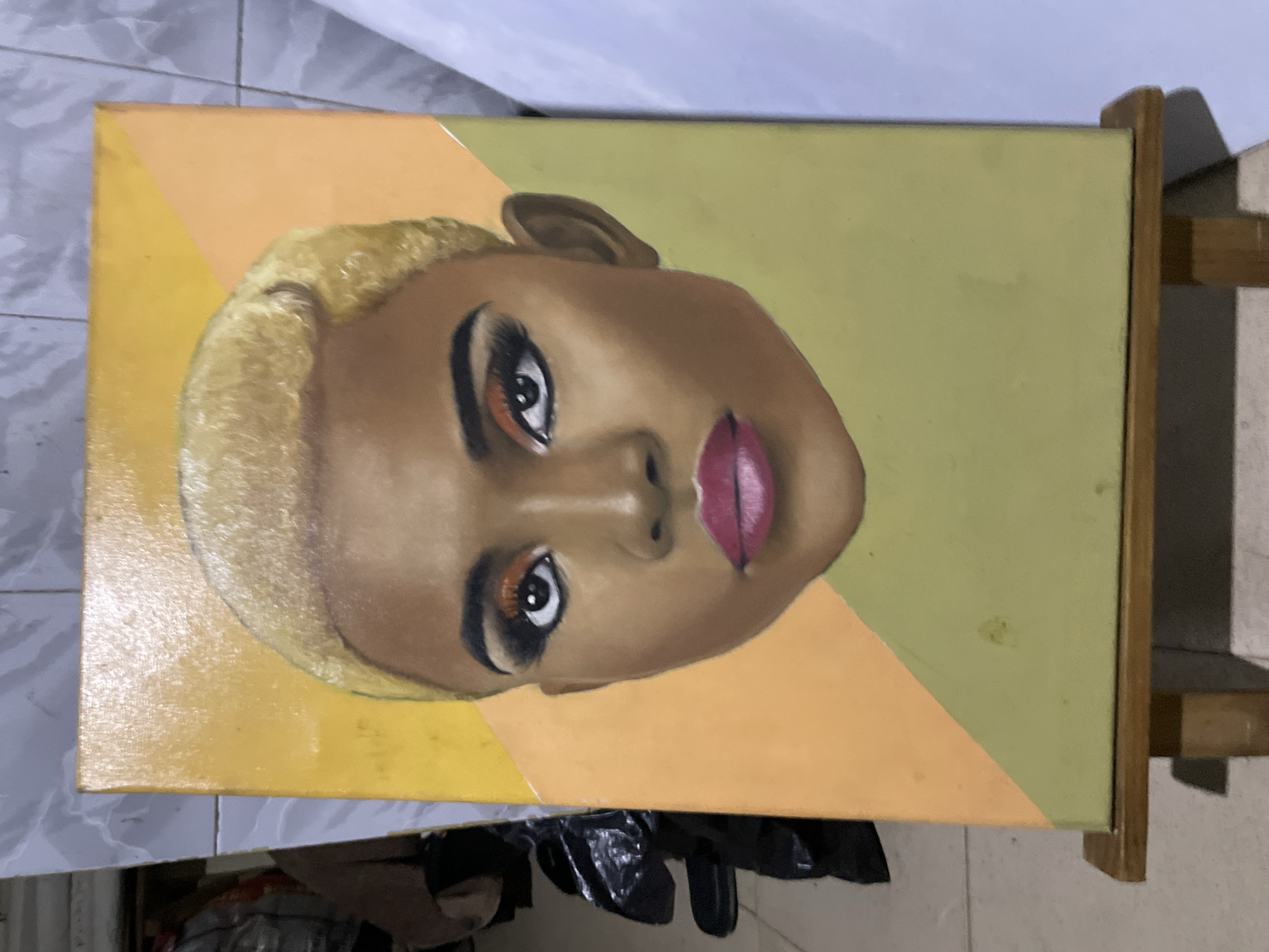 The image shows an oil painting of a woman's face on a canvas, likely situated indoors in Ado, Nigeria. The canvas is partially supported by a wooden easel or stand. The woman in the portrait has fair skin, short blonde hair, and is wearing makeup. The background of the painting is divided into yellow and green sections. In the foreground, there is a glimpse of the surface supporting the easel, along with discarded plastic bags and other objects. The precise street location in Ado, Nigeria cannot be determined from the image.