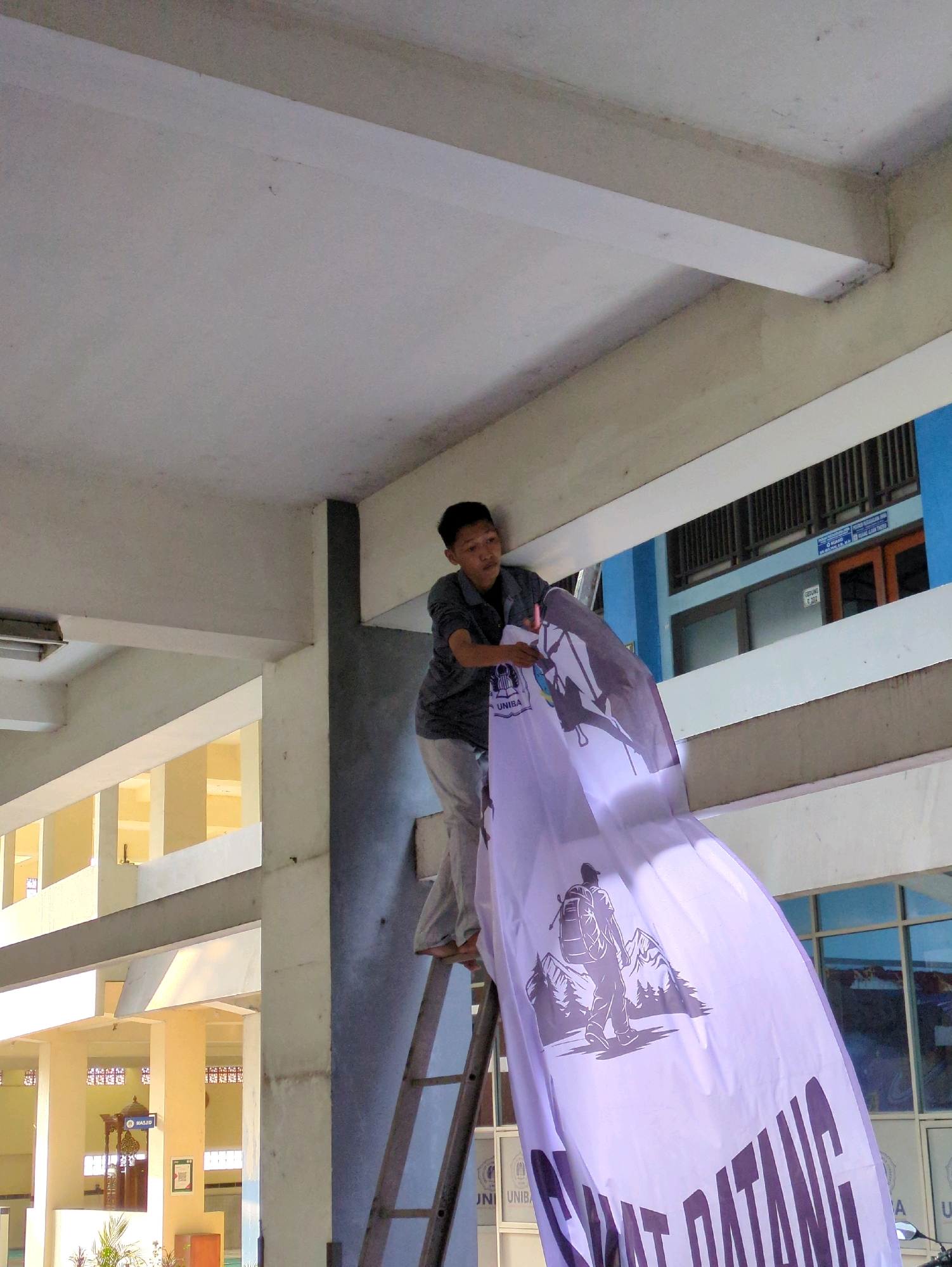 In Surakarta, Indonesia, a young person is positioned on a wooden ladder, actively engaged in hanging a large, light purple banner from a building's overhang. The banner features a graphic of a hiker with a backpack ascending a mountain, rendered in dark purple. Below the graphic, bold, dark purple text reads "SELAMAT DATANG," meaning "Welcome" in Indonesian. The environment appears to be an educational institution or public building, evidenced by the architectural elements such as columns, walkways, and windows. The presence of a mosque sign in the background suggests the location might be part of a university campus or a complex that includes religious facilities. The lighting indicates it is daytime, with sunlight casting shadows and highlighting the building's structure.