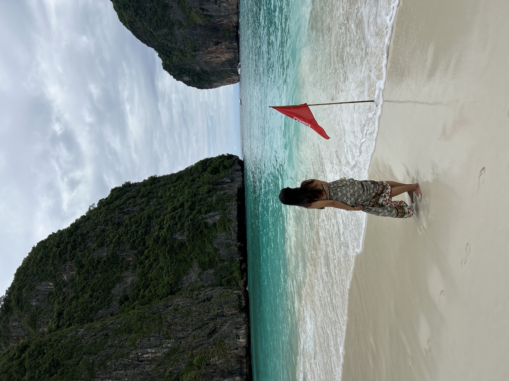 A woman stands on a sandy beach, her back to the camera, facing the turquoise water. She is wearing a patterned sarong. A red flag on a wooden pole is planted in the sand to her right, near the water's edge. The beach is flanked by steep, jungle-covered cliffs. The sky is cloudy. The scene appears to be Maya Bay, Phi Phi Islands, Krabi Province, Thailand, which is a popular tourist destination accessible by boat from Ao Nang.