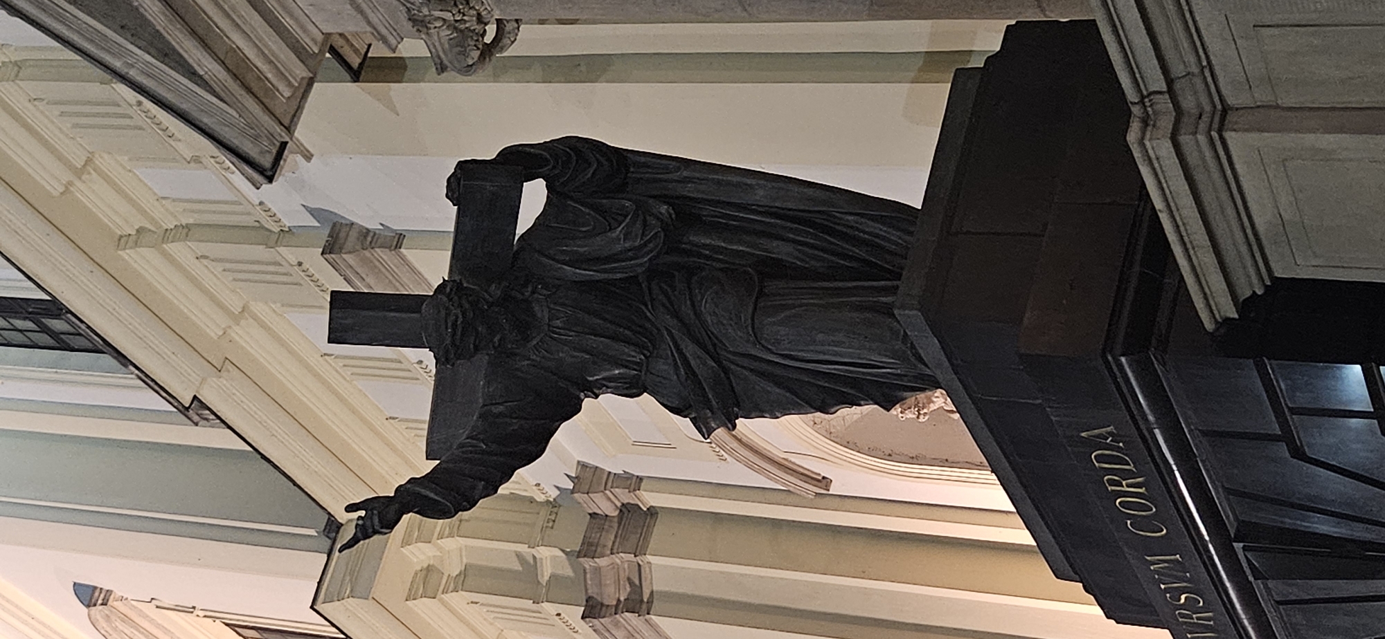 The image shows a dark bronze statue of Jesus Christ carrying a cross, set against the facade of a light-colored building. Jesus is depicted with a crown of thorns, his head bowed, and his right arm raised with fingers pointing upwards. The cross is held over his left shoulder and upper back. The statue is positioned on a dark pedestal, which has text inscribed on its front surface. The text appears to read "IVRSVM CORDA". The building behind the statue has architectural elements like pilasters, cornices, and arched window recesses, suggesting it is a significant historical or religious structure. The lighting suggests it is nighttime or dusk.
This statue is located on the facade of the Archcathedral of St. John the Baptist in Warsaw, Poland. It is one of the sculptures adorning the exterior of the cathedral.