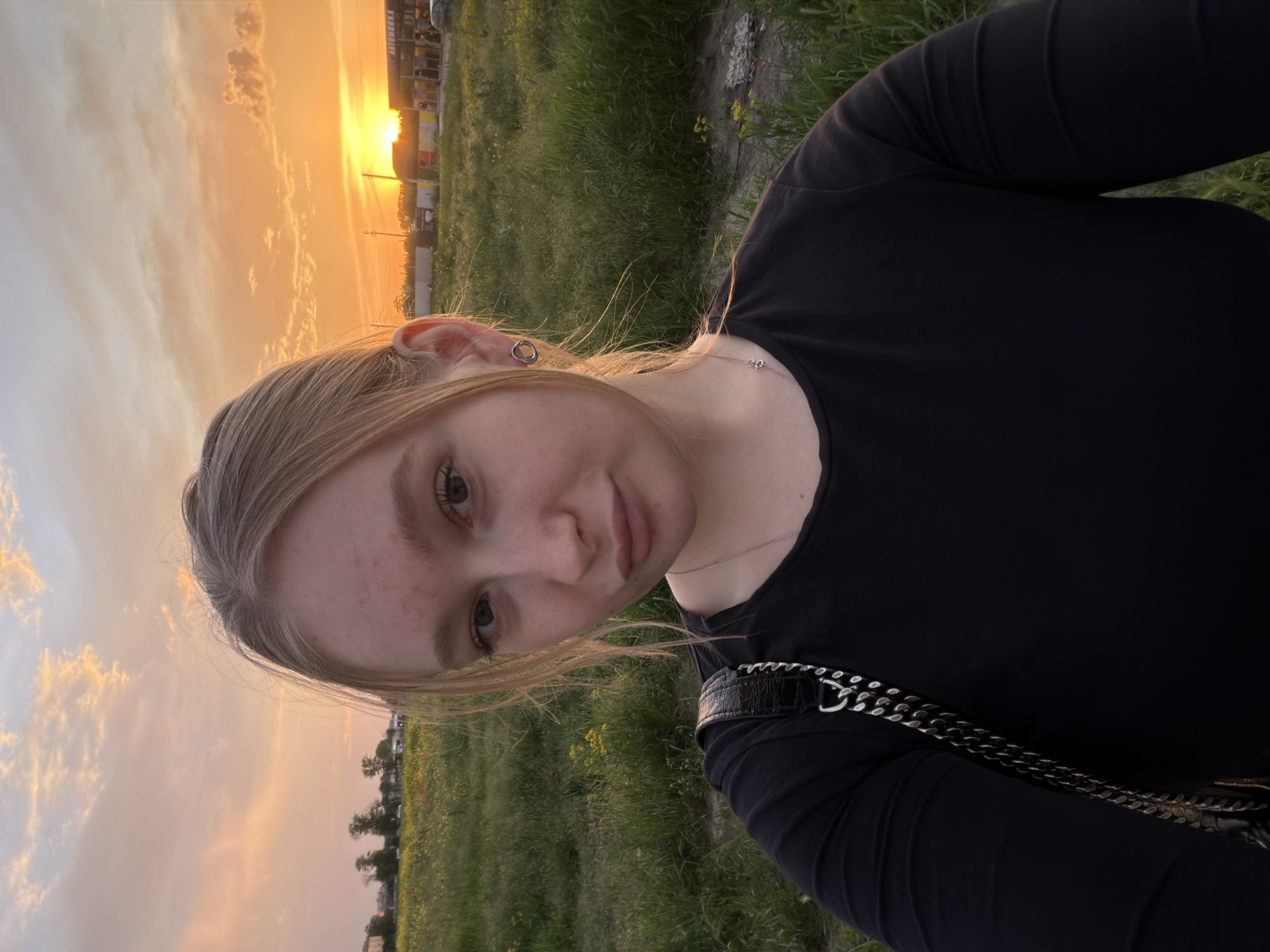 A young woman is taking a selfie outdoors during a sunset. She has fair skin, blonde hair, and is wearing a black long-sleeved shirt and a silver necklace. The background features a field of green grass and buildings under an orange and yellow sky. The location is Krasnodar, Russia.