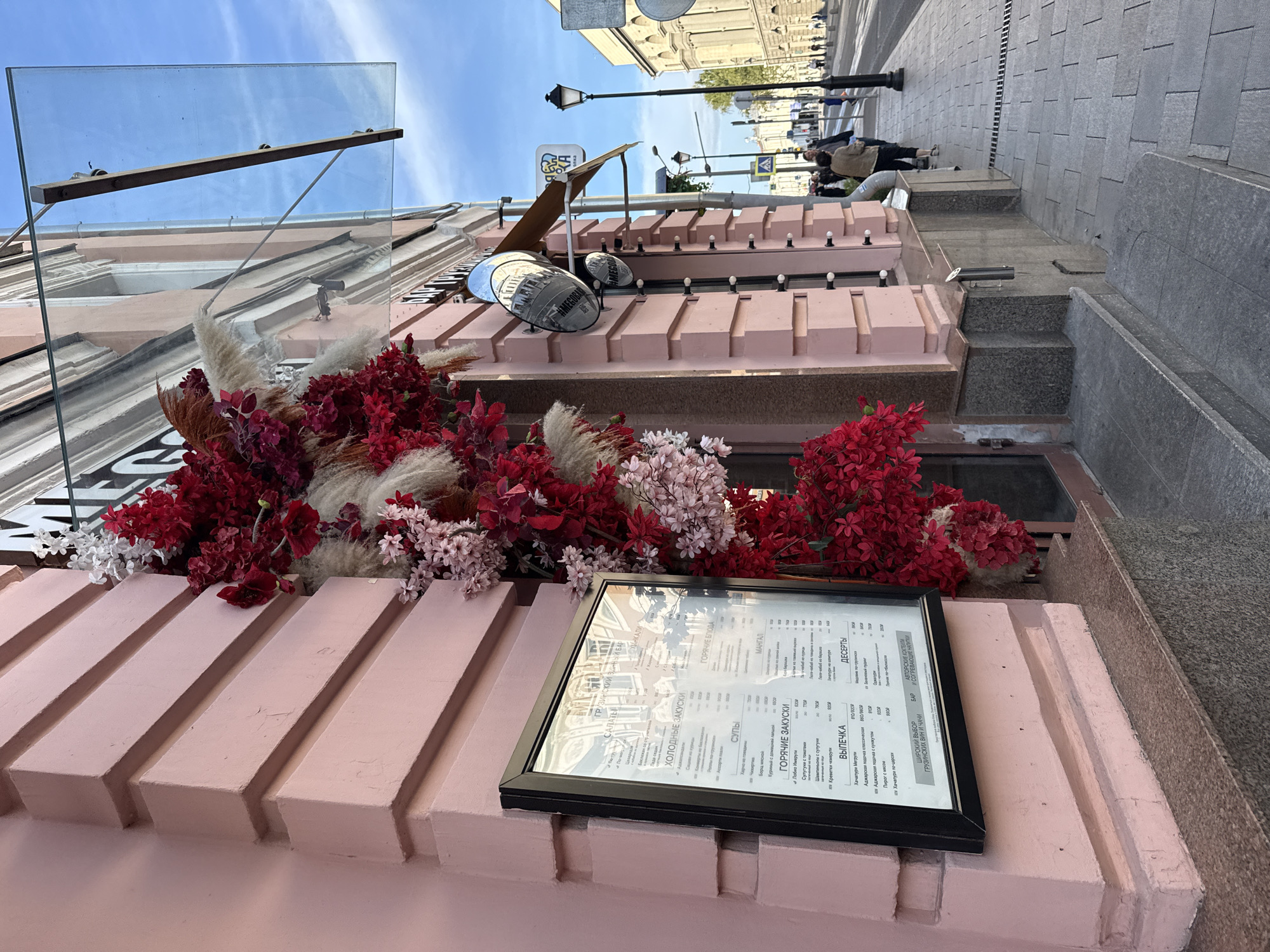 An outdoor, eye-level, medium shot captures the facade of a pink building in Moscow, Russia. The building features a glass awning above its entrance. A large decorative arrangement of red and white flowers and beige plumes cascades down the wall beside a framed menu. The menu, titled "ГРУЗИНСКИЙ ВИННЫЙ БАР" (Georgian Wine Bar), displays various food categories such as "ХОЛОДНЫЕ ЗАКУСКИ" (cold appetizers), "ГОРЯЧИЕ ЗАКУСКИ" (hot appetizers), "СУПЫ" (soups), and "ВЫПЕЧКА" (pastries). To the right of the entrance, a street with pedestrian and vehicle traffic is visible. A lamppost stands on the sidewalk, which is paved with gray stones. Several pedestrians are walking along the street, and cars are visible in the distance. The sky is blue with some clouds.