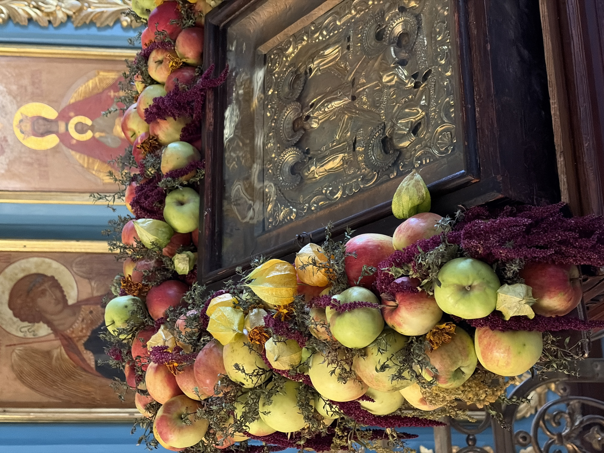 The image depicts an Orthodox iconostasis, a wall of icons and religious images, likely within a church or religious setting in Moscow, Russia. The icons are framed and ornate, with gold halos. In the foreground, a decorative arrangement of fruits and foliage adorns the front of the iconostasis. The fruits appear to be a mix of apples and possibly physalis, interspersed with dark purple amaranth or similar floral elements and dried greenery. The overall impression is of a devotional display, possibly for a religious festival or commemoration. The lighting is soft, suggesting an indoor environment.