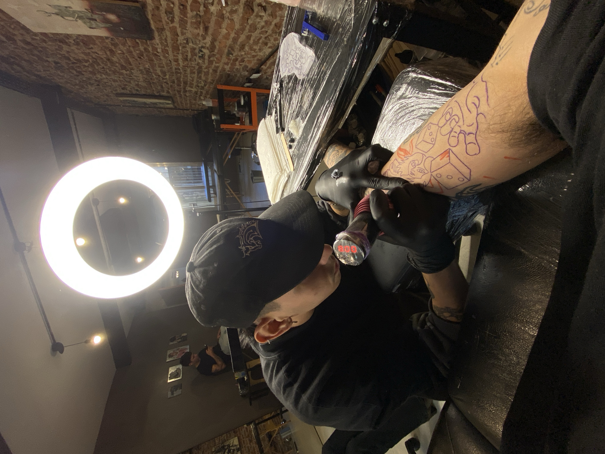 The image depicts a tattoo artist meticulously working on a client's forearm inside what appears to be a tattoo studio in Buenos Aires, Argentina. The artist, positioned on the left side of the frame, is wearing a black cap with a dark, intricate logo, a black long-sleeved shirt, and black gloves. They are deeply focused on the tattooing process, holding a tattoo machine with a clear grip that faintly displays red text, possibly "880" or "B80".
A bright, circular ring light illuminates the immediate workspace, highlighting the artist's hands and the client's arm. The client's forearm, which is visibly hairy and already bears other tattoos, has a new purple stencil outline of various cartoonish designs, including dice and abstract shapes, ready for inking. Some red marks are also visible on the skin.
The workstation directly in front of the artist is covered in clear protective plastic wrap, a standard hygiene practice in tattoo parlors. On this surface, supplies like paper towels, small ink cups, and needle cartridges are neatly arranged. Another stencil design on white paper lies on the table. In the background, there's a rustic brick wall adorned with framed artwork, and a darker wall with additional pictures. Another individual can be seen in the background, seated and appearing to look at a phone. The overall setting is professional and sterile, typical of a tattoo parlor environment.