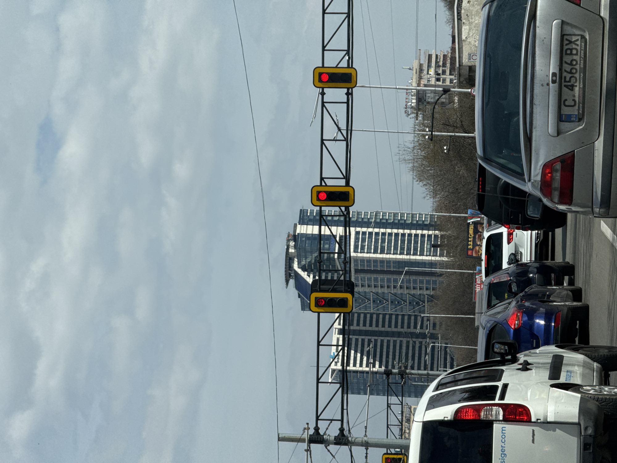 The image depicts a static vehicular traffic scene on a multi-lane road in Sofia, Bulgaria, with prominent high-rise buildings in the background. The location is identified as the vicinity of Tsarigradsko Shose Boulevard, facing towards the **Capital Fort** building, which is visibly located at Tsarigradsko Shose 90.
In the foreground, a column of vehicles is stopped or moving slowly. On the far left, the rear of a white commercial van, displaying "siger.com" on its tailgate, occupies the leftmost visible lane. Immediately behind it, a blue hatchback is visible. In the adjacent lane to the right, a silver sedan with Bulgarian license plate **C.4566 BX** is prominently featured, showing some dirt accumulation on its rear bumper and trunk. Behind this sedan, a white SUV is partially visible. An overhead gantry across the road supports three rectangular traffic lights, all illuminated red, indicating a stop for the vehicles. Overhead wires and utility poles are present.
The midground features a billboard on the right, partially obscured, advertising "M 3.2.1" with images of food, likely hamburgers. A circular speed limit sign, possibly "70", and a "ЗОНА" (ZONE) sign are also visible on a pole to the right. Bare deciduous trees are situated between the road and the background structures.
The background is dominated by modern high-rise architecture under an overcast sky with patches of lighter clouds. The most prominent structure is the **Capital Fort** building, characterized by its distinctive sloped, multi-faceted roof with what appears to be a helipad and reflective surfaces, situated to the right of the center. To its left, another tall, contemporary office building with alternating light and dark horizontal bands is visible. Additional, less distinct buildings can be seen further in the distance. No individuals are clearly discernible in the image.