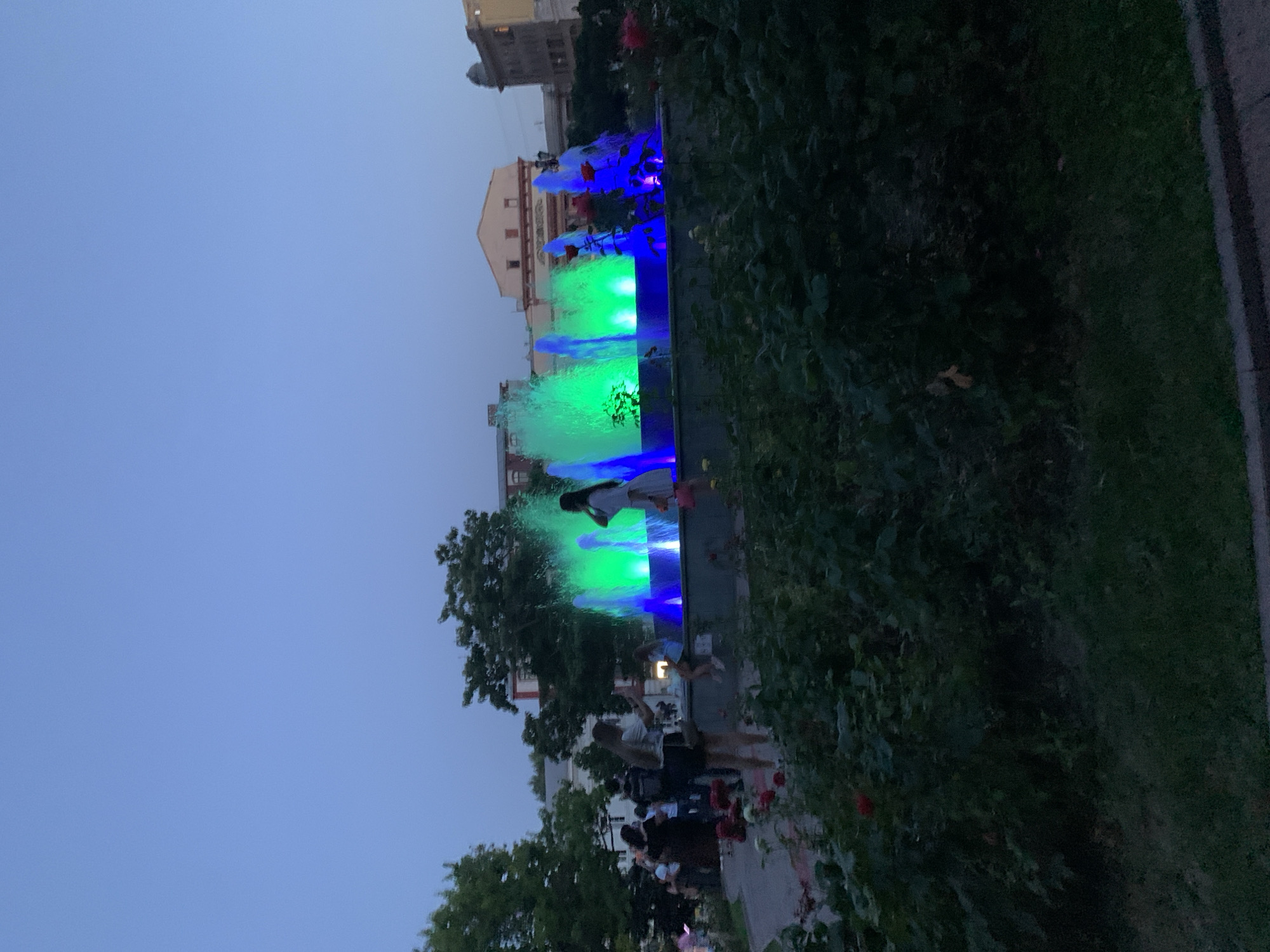 The image depicts a public park scene in Odesa, Ukraine, during twilight. A large, illuminated fountain is the central focus, with water jets lit in vibrant green and blue hues. Several people are visible in the background, some near the fountain and others standing in the park. In the foreground, lush green bushes and some red roses are present, suggesting a well-maintained garden area. The sky is a clear, deep blue, characteristic of the evening hour. Buildings, likely in the surrounding urban area, are visible in the distance behind the fountain and trees. The overall atmosphere is one of a leisurely evening gathering in a city park.