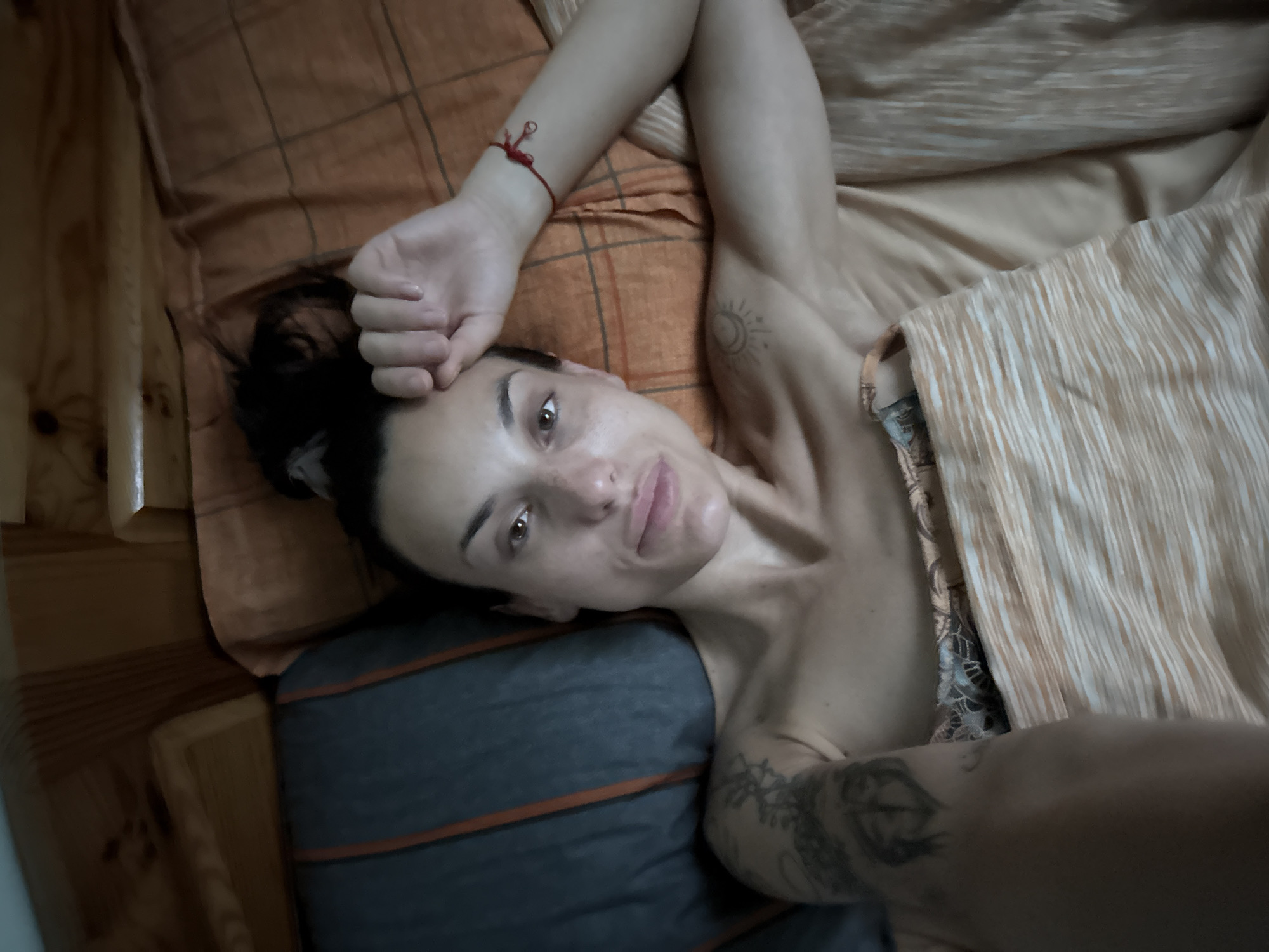 The image shows a woman lying in a bed. She is looking at the camera and has her hand raised to her forehead. She has dark hair pulled up in a bun and is wearing a red bracelet on her wrist. She has tattoos on her left arm and shoulder. The bed appears to be in a room with wooden walls and orange and gray bedding. There is no indication of the location in the city of Pleven, Bulgaria.