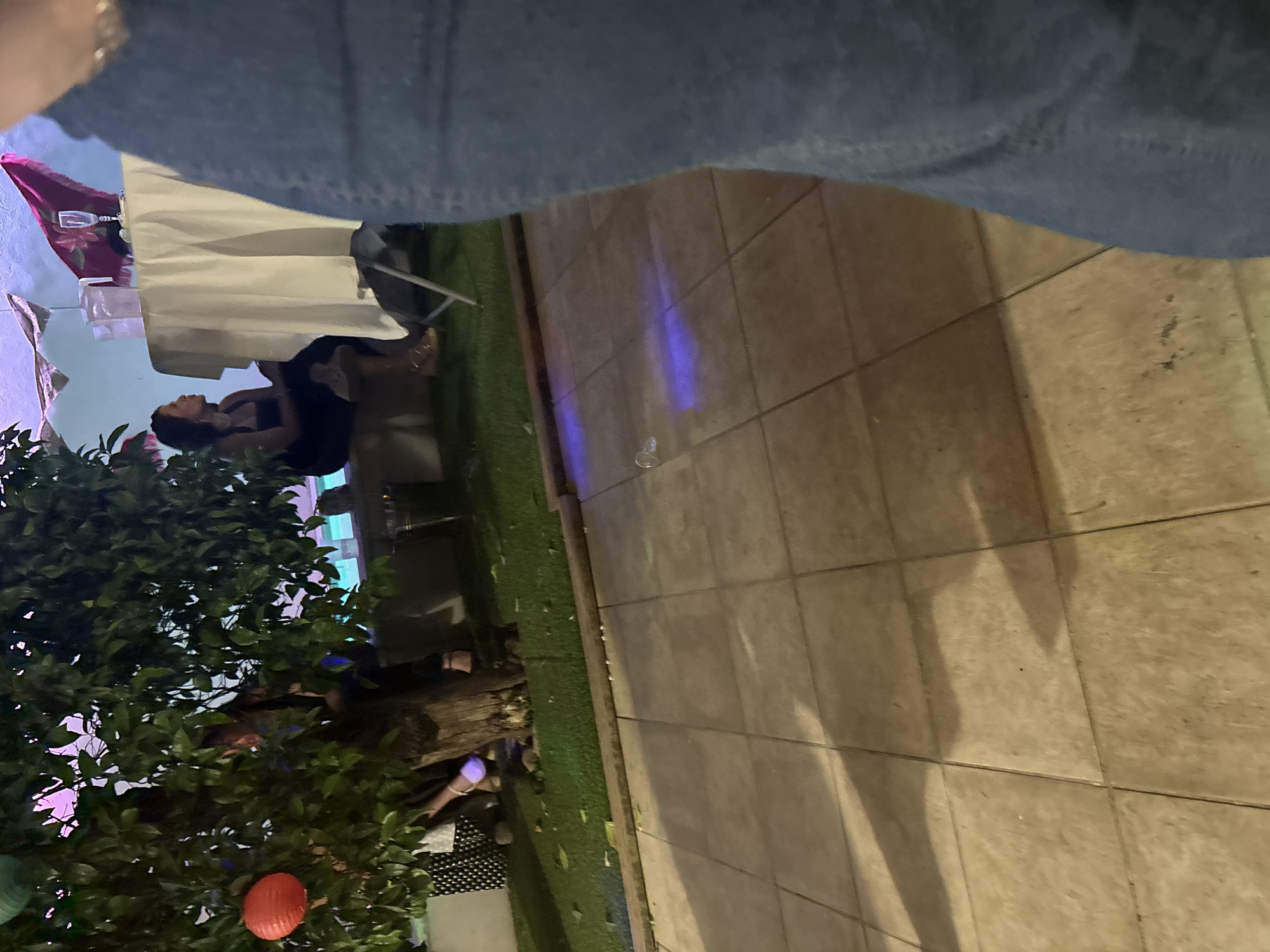 The image shows an outdoor patio setting, likely in Córdoba, Spain. A group of people are seated at a table covered with a white tablecloth, with a red object, and possible dining items visible. A large tree with a red lantern hanging from it is in the foreground. A paved area with shadows is to the right. A person's leg is visible in the top part of the frame. The lighting suggests it is evening.