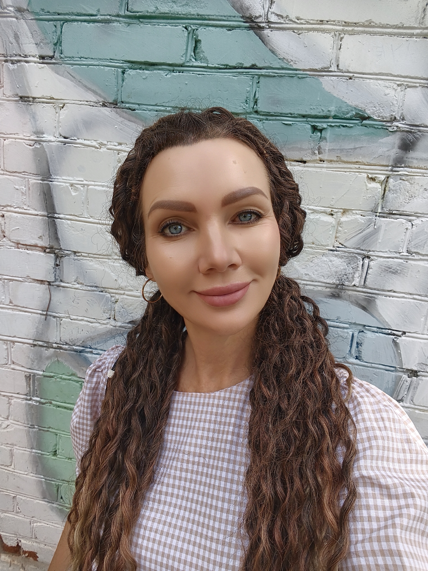 A woman with long, curly brown hair braided into two pigtails stands in front of a white brick wall with a light blue and gray graffiti design. She is looking at the camera and smiling. She is wearing a light brown and white checkered dress. The photo appears to be a selfie.