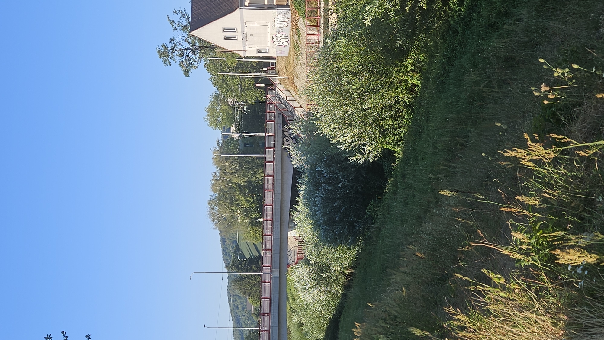 The image depicts a sunny day scene, likely in Prešov, Slovakia, featuring a modern bridge spanning a body of water, possibly a river or stream. The bridge is constructed of concrete and has red railings along its sides. Vegetation, including lush green bushes and tall grasses, flanks the watercourse and slopes upwards towards the bridge.
On the right side of the image, a light-colored building with a dark roof is visible. The building has windows and is adorned with graffiti. Adjacent to the building, a set of stairs with red railings leads up towards the bridge level. In the background, a tree-lined hill or small mountain rises, with trees and power lines visible in the mid-ground. The sky is clear and blue.
The image appears to be taken from a slightly elevated position, looking towards the bridge and the building. There are no people visible in the frame. The overall impression is of a semi-urban or suburban landscape during daylight hours, with the bridge serving as a significant structural element connecting different parts of the area. The graffiti on the building and the presence of power lines suggest an inhabited area.